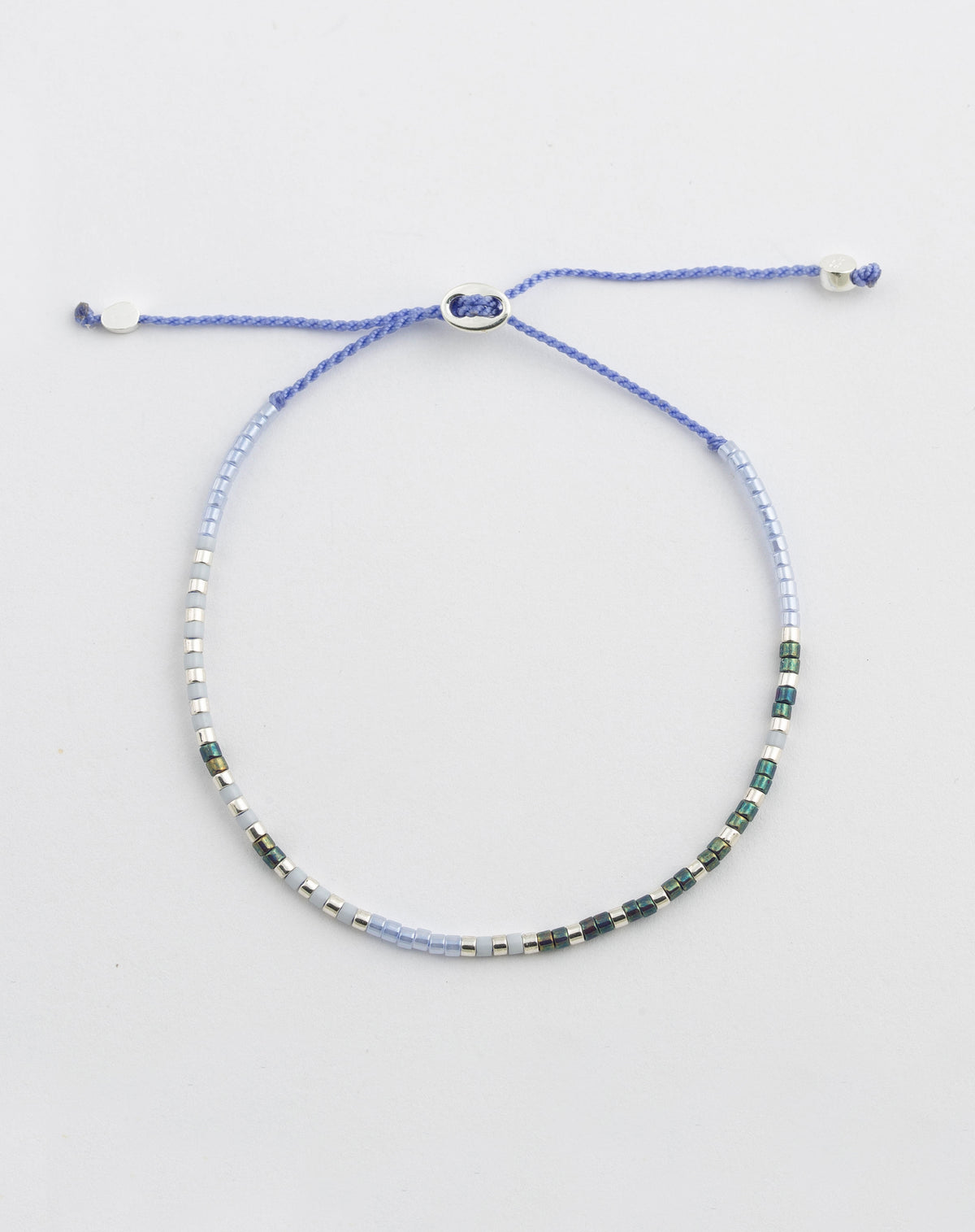 A product shot of the Syster P Code Bracelet in the "Good Vibes" style, showing the purple cord adorned with small silver, light blue, and dark green glass beads that spell out the message in morse code.