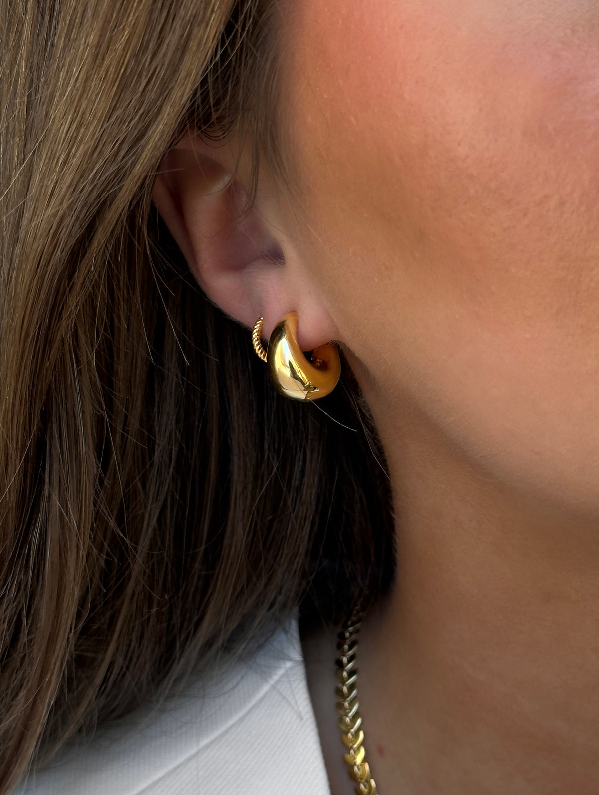 A close-up shot of a model wearing the Bolded Little Sis Earrings in gold, featuring a chunky hoop design with a twisted detail at the clasp, and a matching gold chain necklace.