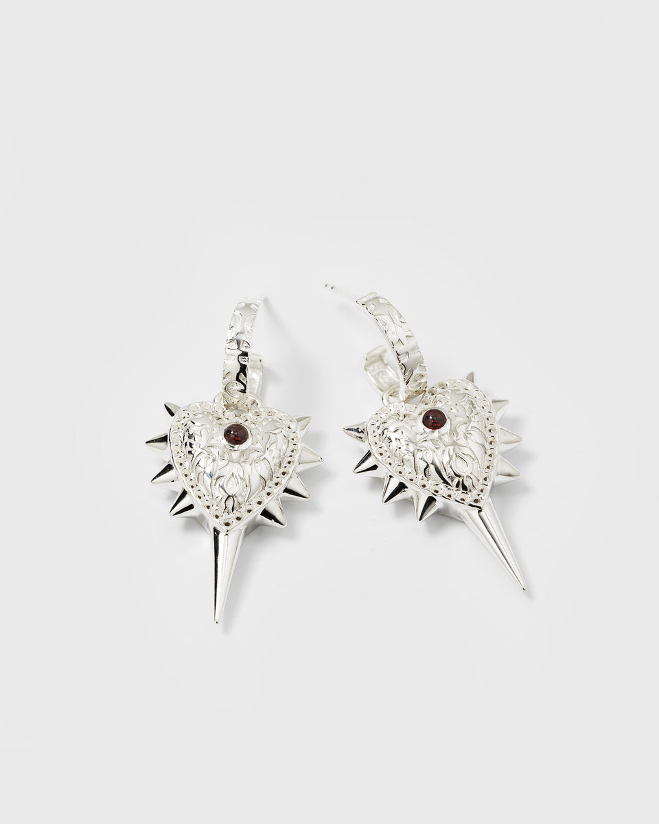 A product shot of the Drama Queen Heart Spike Earrings in silver, featuring patterned hoops with dangling heart charms adorned with spikes and a central red stone.