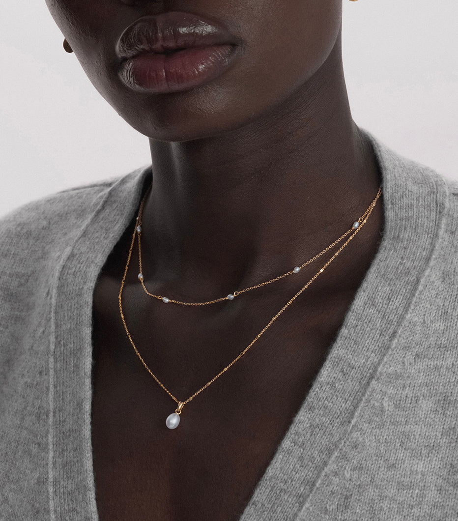 A close-up of a model wearing the Treasure Single Pearl Necklace in gold, featuring a delicate teardrop-shaped pearl, layered with a shorter gold necklace dotted with small freshwater pearls.