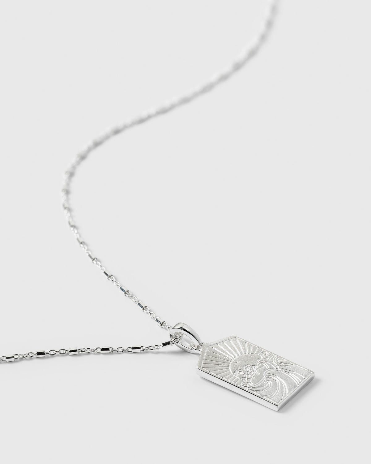 A close-up product shot of the Lucky Coin Keep Dreaming Necklace in silver, highlighting its rectangular pendant which is engraved with a detailed paradise scene featuring a sun and waves.