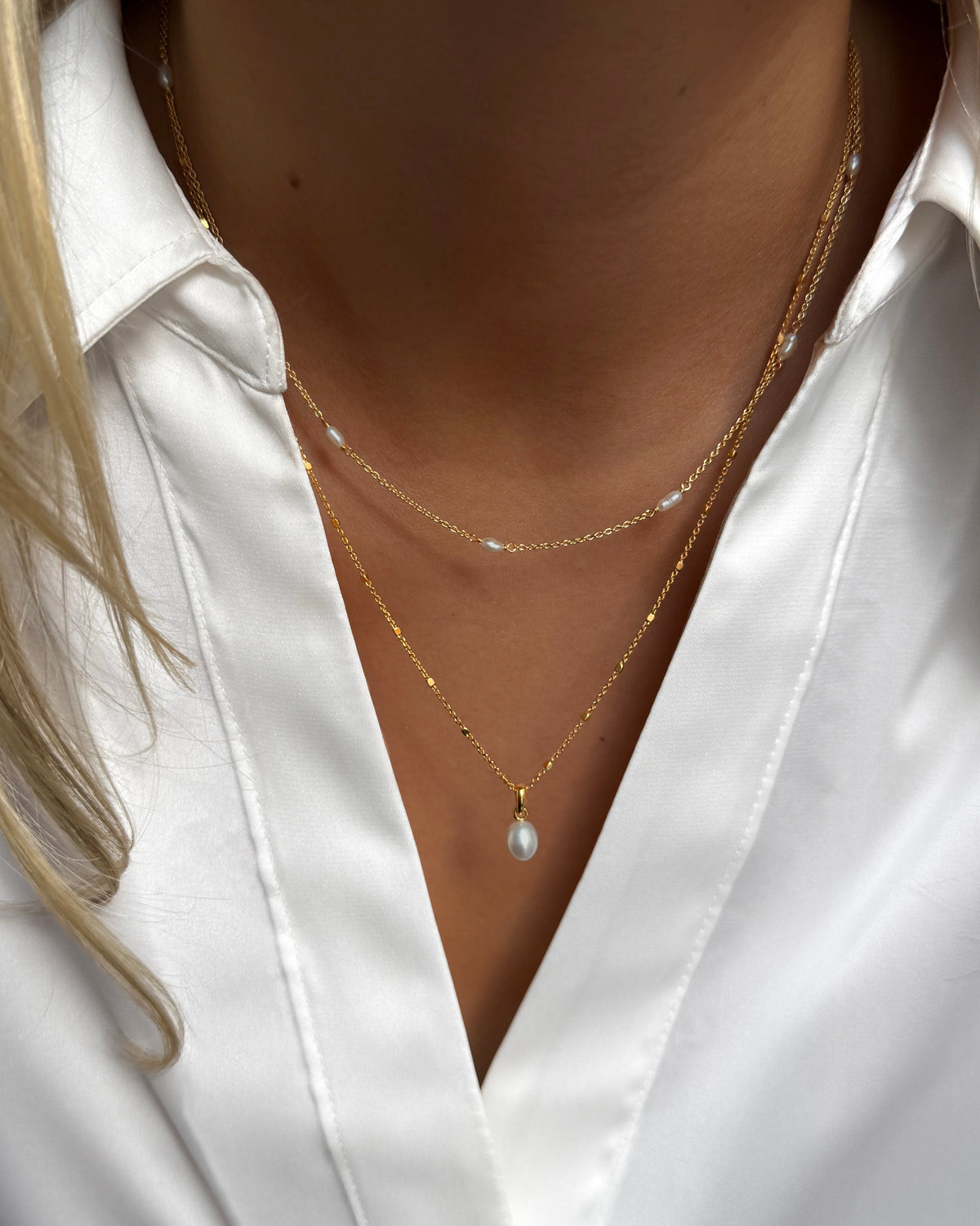 A model wears the gold Treasure Multi Pearl Necklace, styled in a layered look with the Treasure Single Pearl Necklace, both featuring delicate freshwater pearls and shown against a crisp white shirt.
