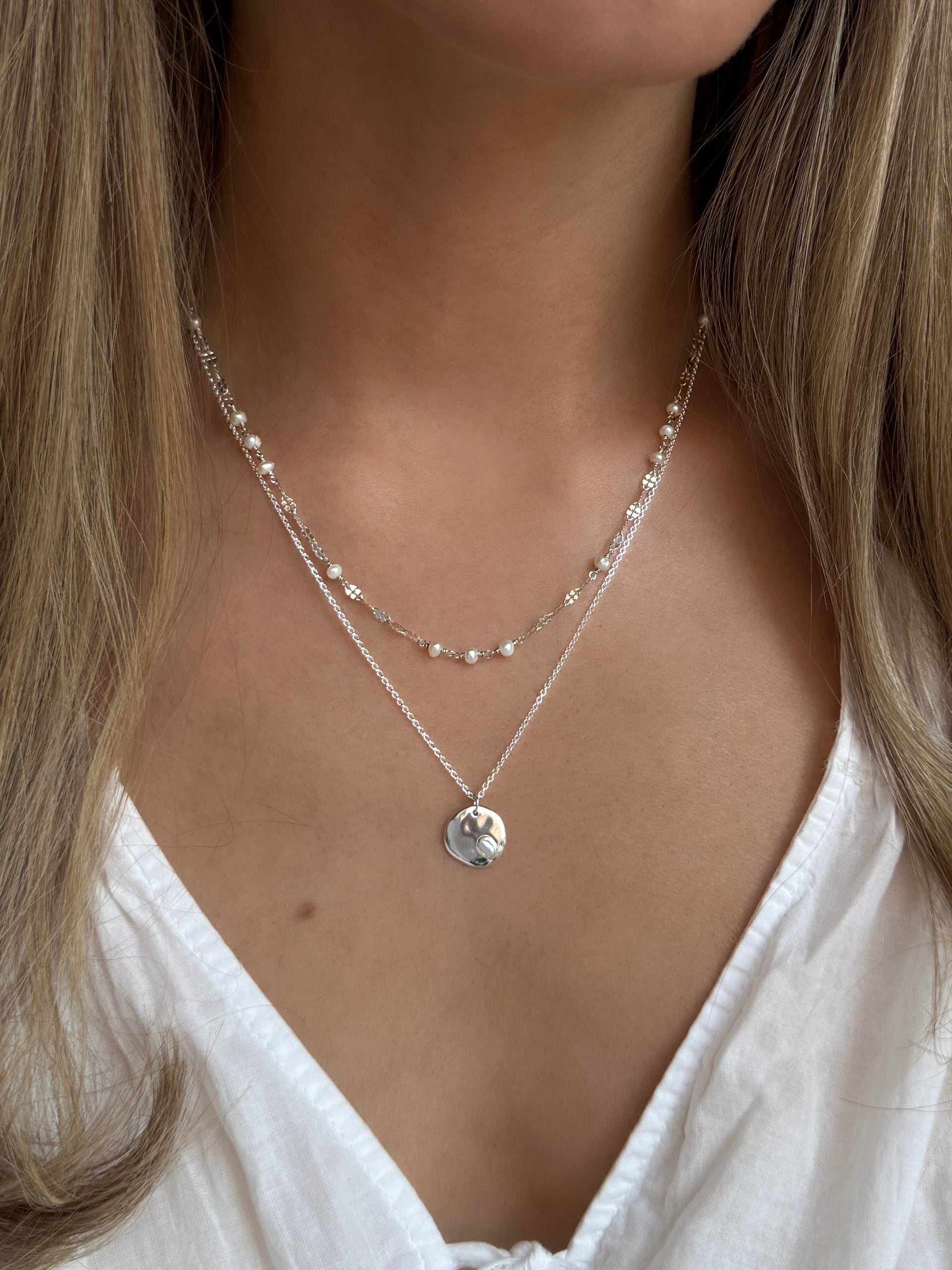 A model wears the silver Aurelia Necklace, featuring a delicate chain with small freshwater pearls, layered with another silver necklace that has a simple chain and a round pendant with a single pearl.