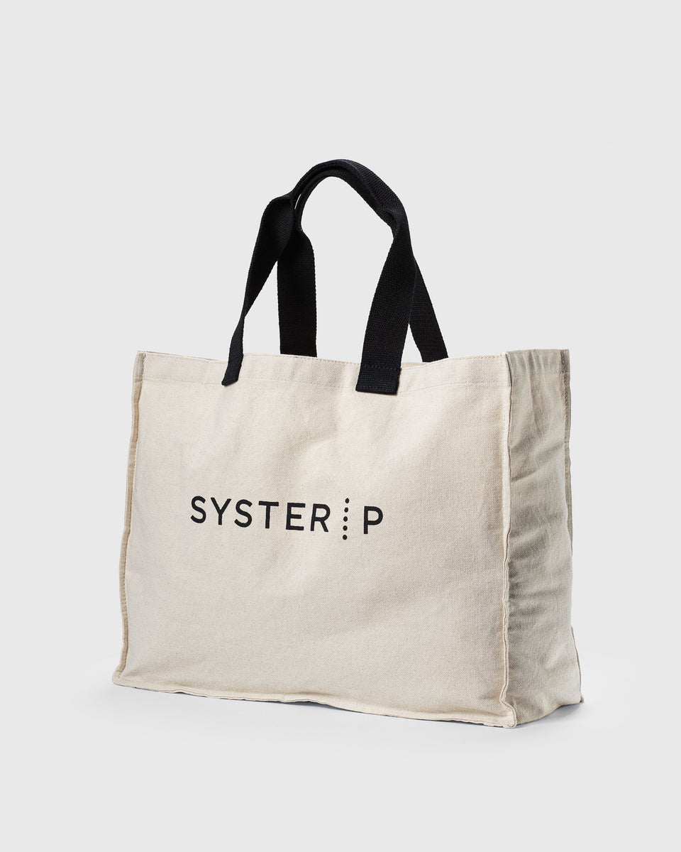 A product shot of the Syster P tote bag, a beige cotton bag with black handles and the brand's logo printed in black on the front.