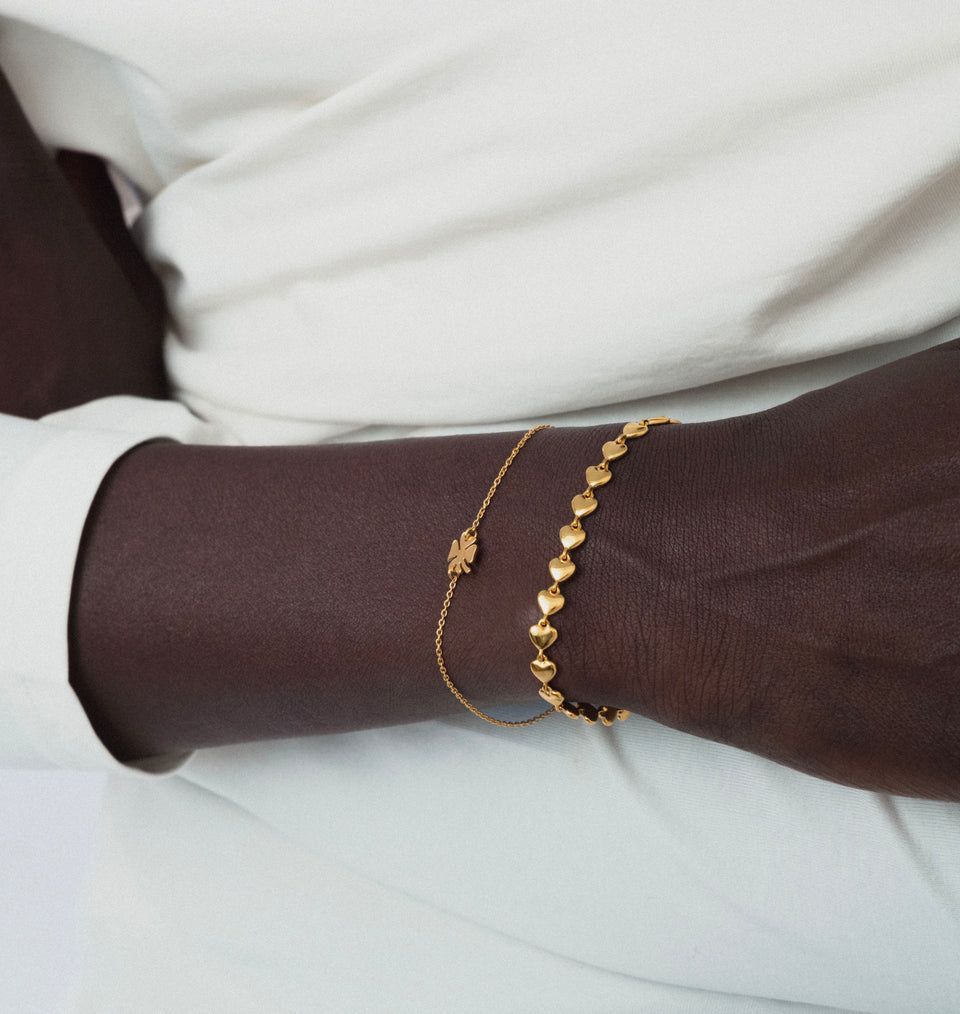 A close-up of a model's wrist wearing the Bring Me Luck Bracelet in gold, featuring a delicate chain and a four-leaf clover charm, styled with a second gold bracelet made of linked hearts.