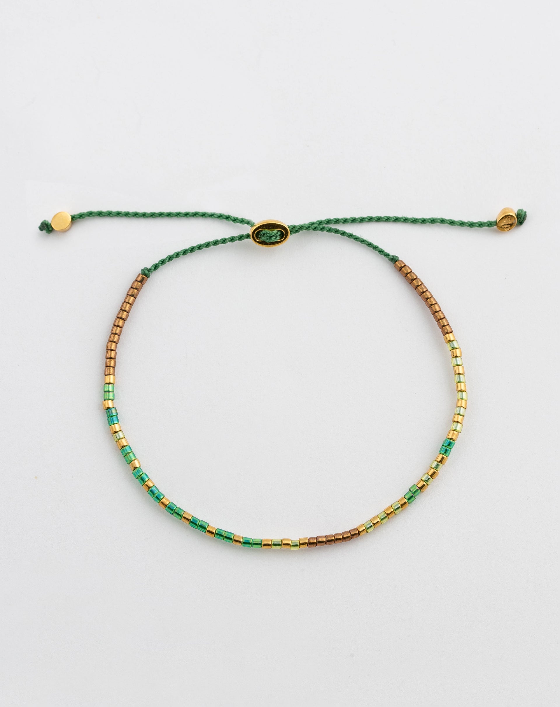 A product shot of the Code Bracelet Gold Good Vibes, laid out in a circle. The bracelet features a green cord and small Japanese glass beads in shades of green, gold, and bronze, which spell out 'Good Vibes' in Morse code.