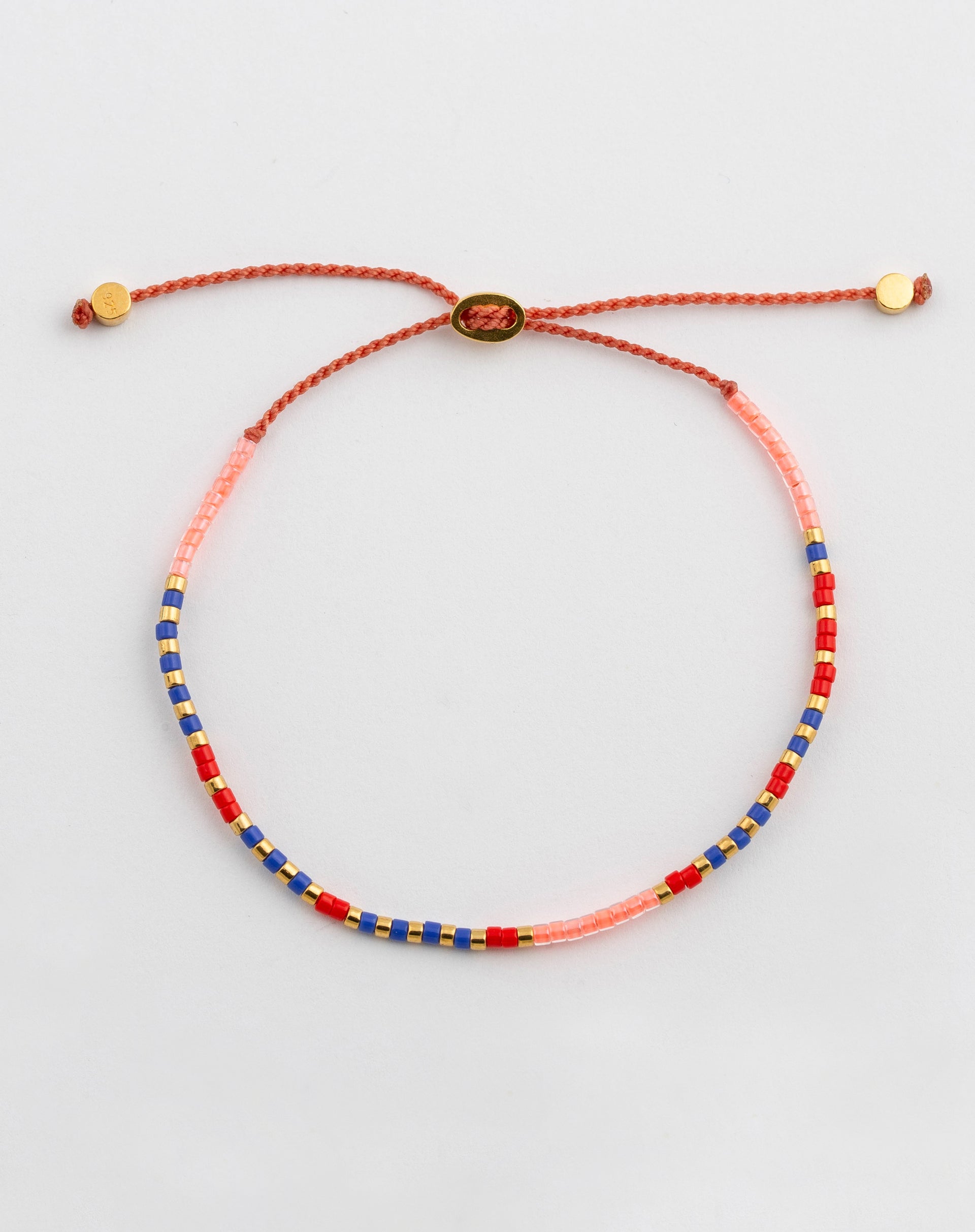 A product shot of the Code Bracelet Gold 'Just Breathe', laid out to display its multicolored beads in pink, red, blue, and gold, which spell out the message in Morse code on an adjustable cord.