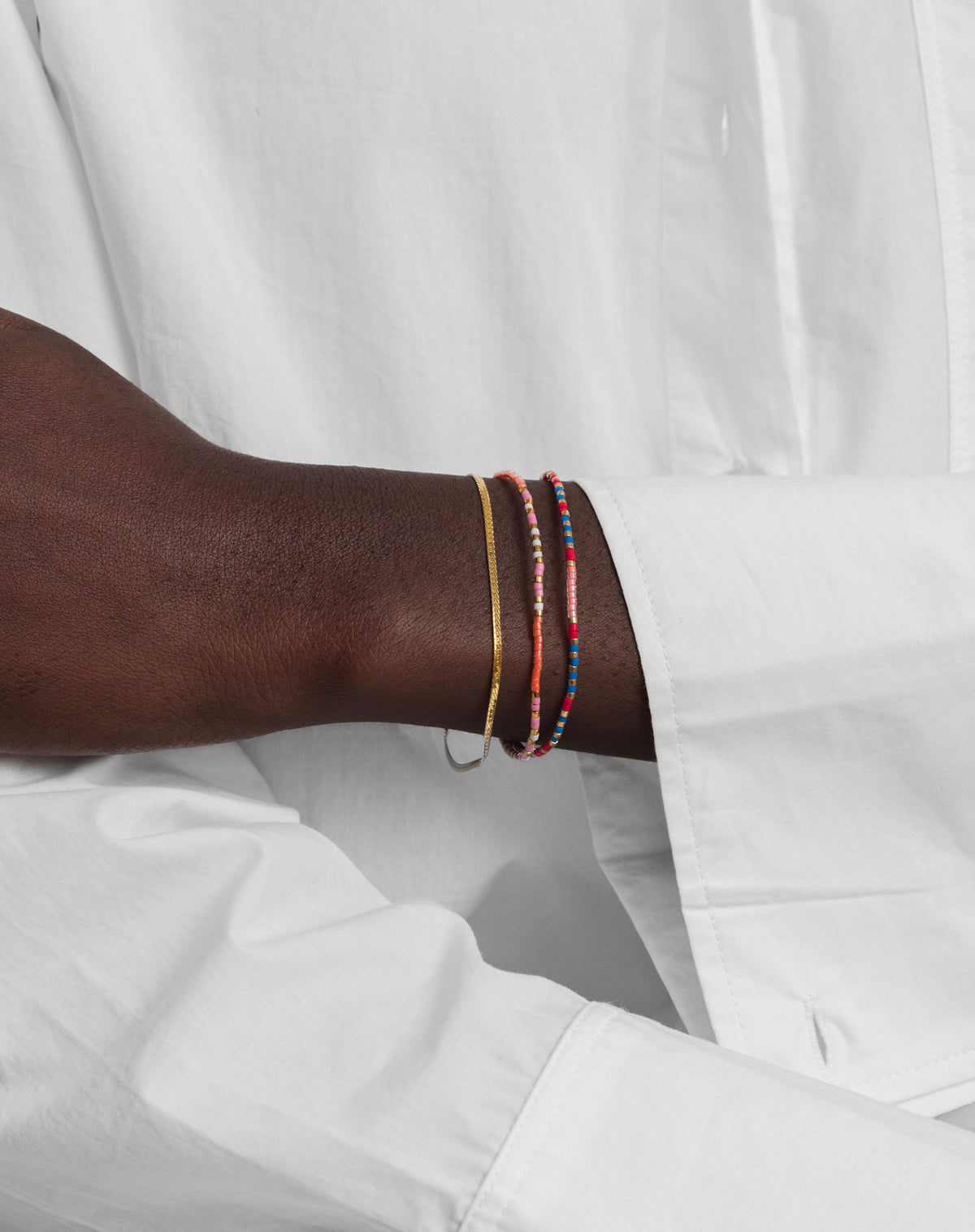 A close-up of a model's wrist, styled with the Code Bracelet Gold 'Just Breathe' alongside another colorful beaded bracelet and a thin gold chain.