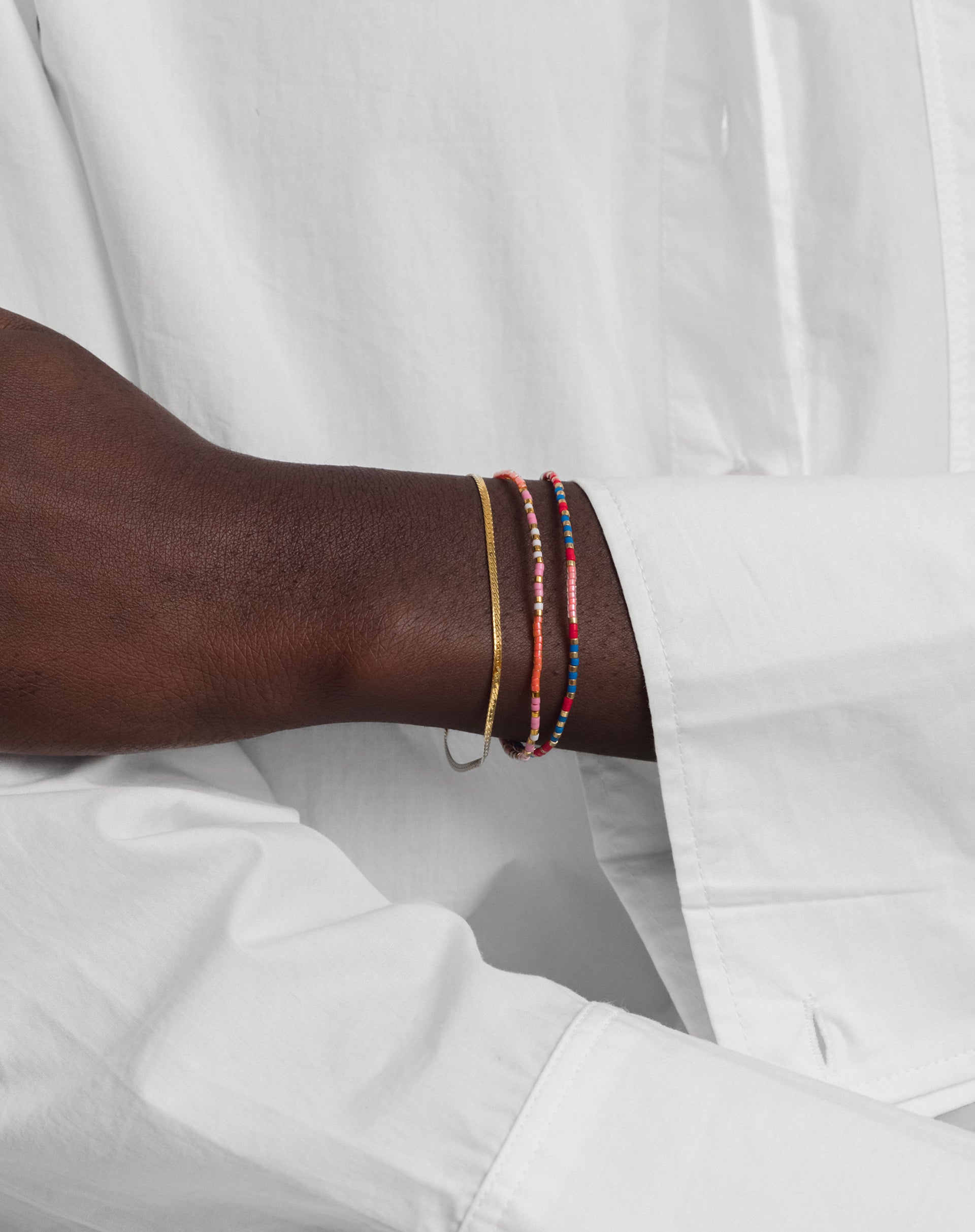 A close-up of a model's wrist, styled with the Code Bracelet Gold 'Just Breathe' alongside another colorful beaded bracelet and a thin gold chain.