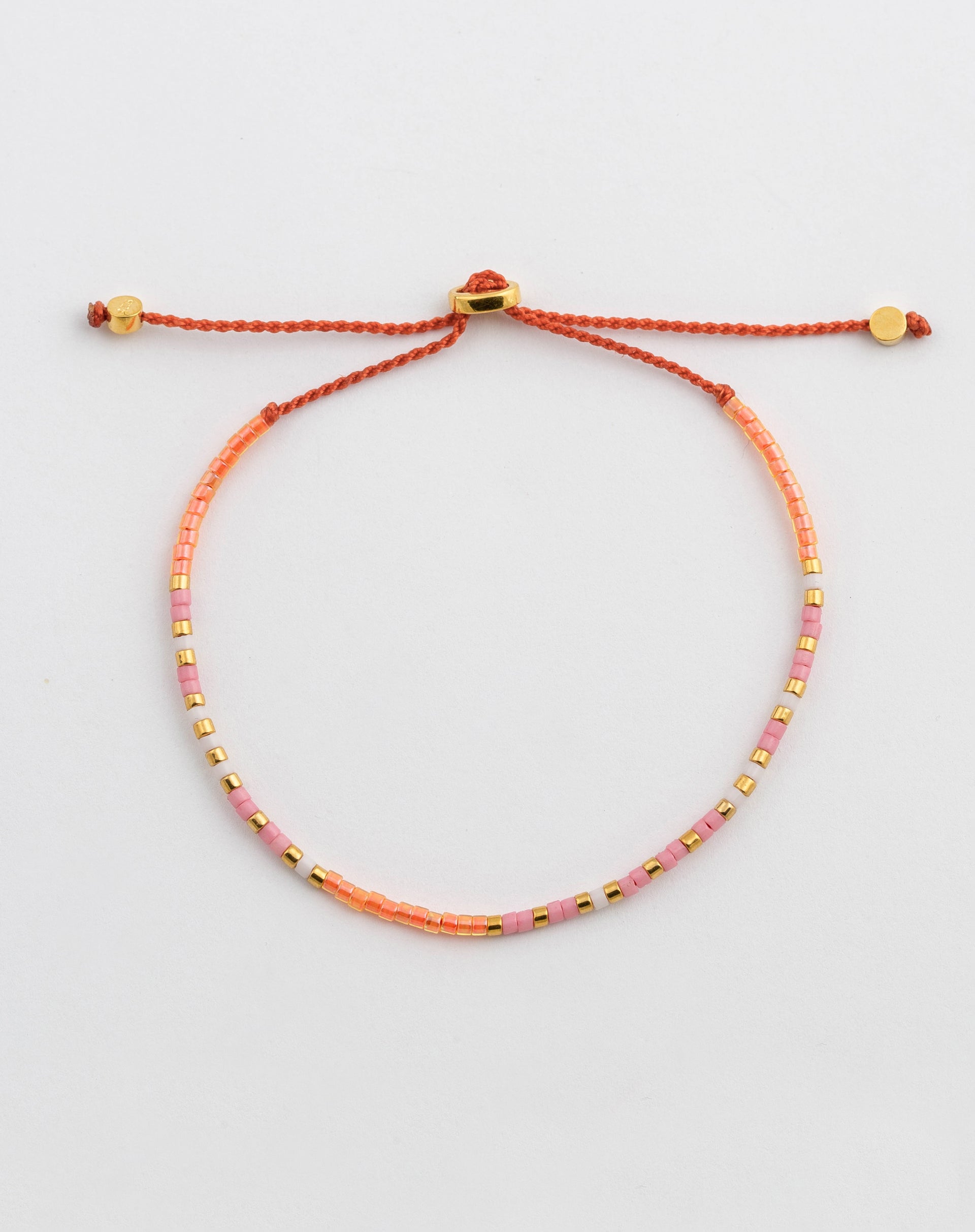 A product shot of the Code Bracelet Gold Keep Going, laid out in a circle to display its colorful pattern of pink, orange, white, and gold beads on an orange cord with a gold-plated adjustable clasp.