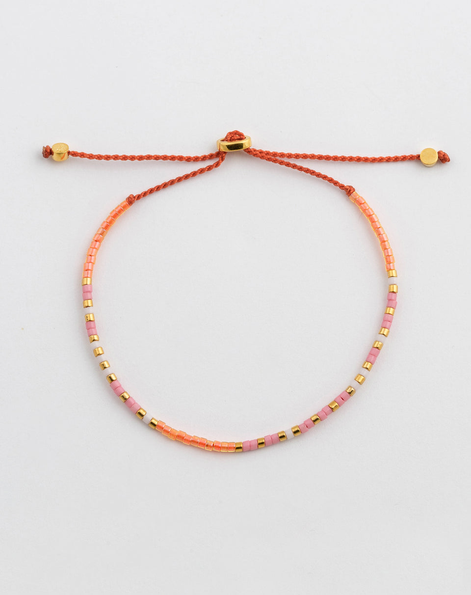 A product shot of the Code Bracelet Gold Keep Going, laid out in a circle to display its colorful pattern of pink, orange, white, and gold beads on an orange cord with a gold-plated adjustable clasp.