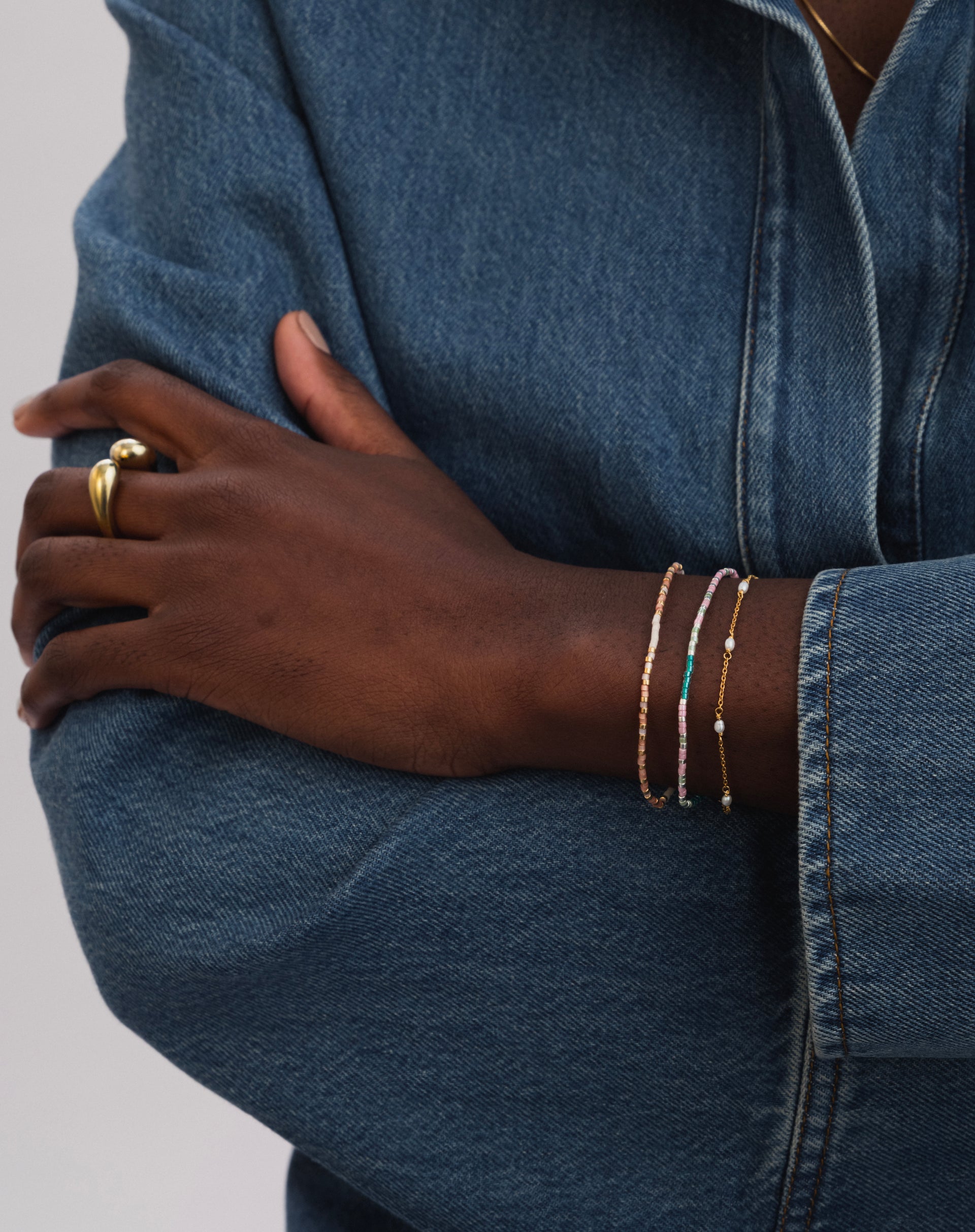 A model wears the Code Bracelet Gold 'Stay Strong', featuring pink and white beads, stacked with another colourful beaded bracelet and a fine gold and pearl chain bracelet. A chunky gold ring is also visible on her finger.