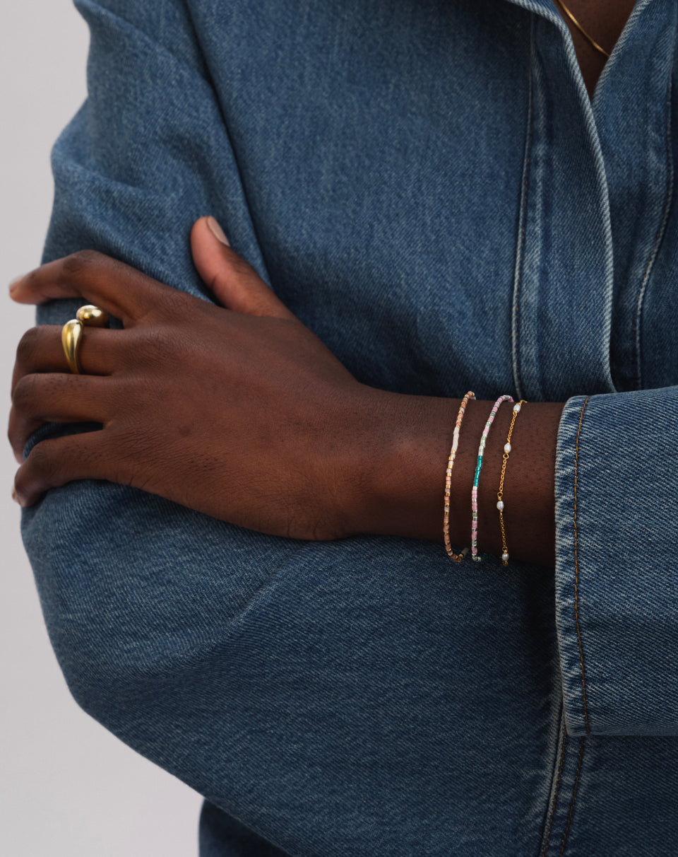 A model wears the Code Bracelet Gold 'Stay Strong', featuring pink and white beads, stacked with another colourful beaded bracelet and a fine gold and pearl chain bracelet. A chunky gold ring is also visible on her finger.