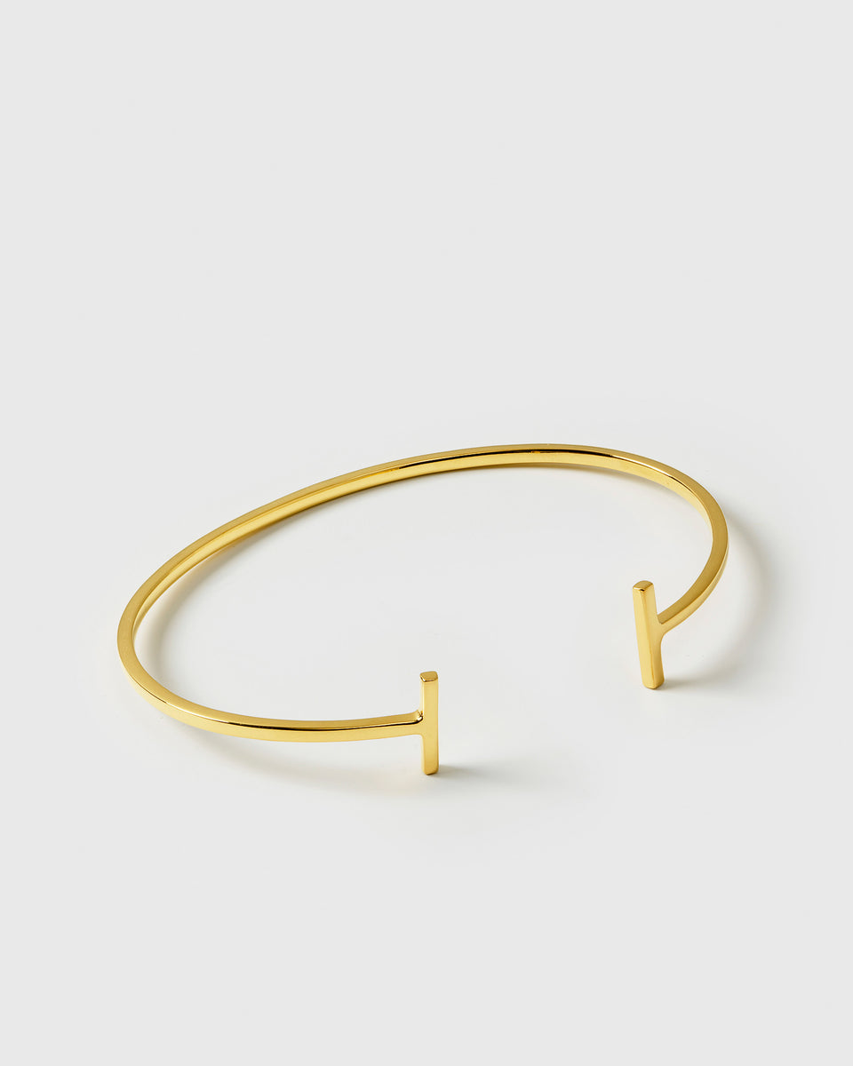 A product shot of the Strict Bar Bangle in gold, showcasing its thin, open design with a geometric bar at each end.