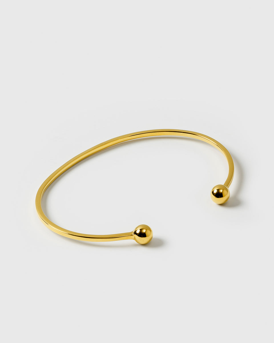 A product shot of the Strict Ball Bangle in gold, showing its thin open cuff design with a small polished sphere at each end.