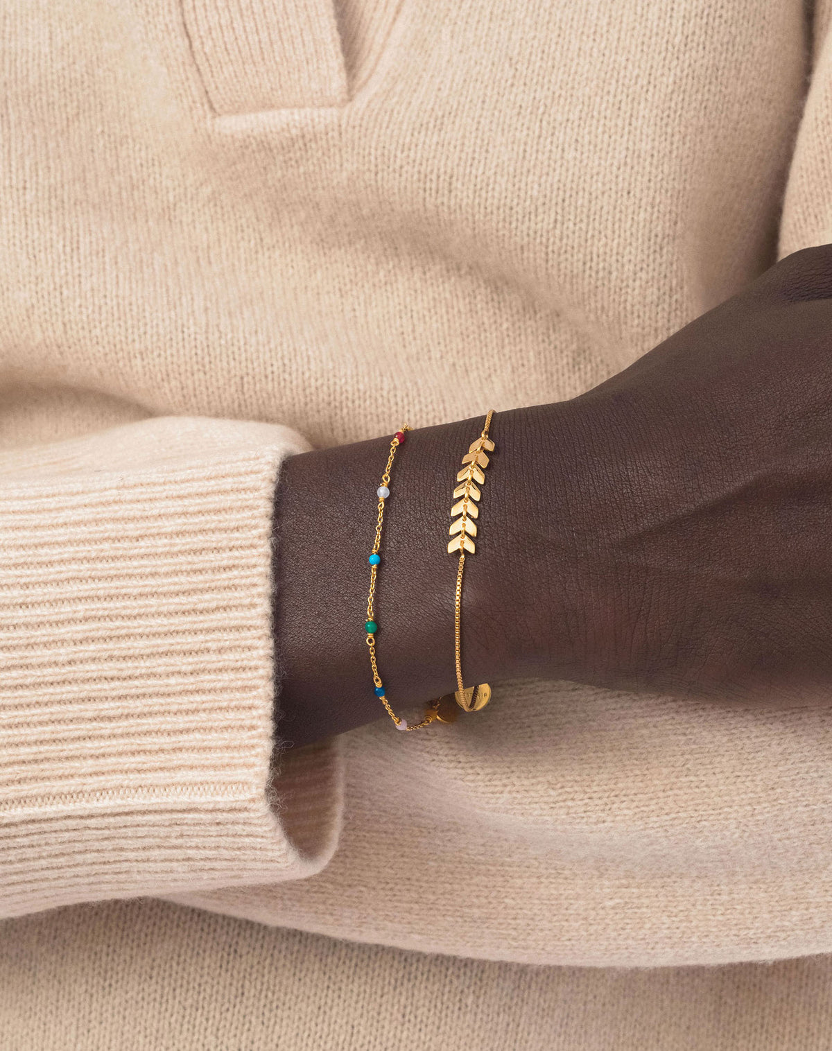 A close-up of a model's wrist, showing the Santa Monica Bracelet Gold with its delicate, multicoloured stones: Yellow Jade, Pink Opal, Sodalite, Green Onyx, Blue Color Reconstituted Turquoise, Vit Agate and Carnelian. Layered with another gold bracelet designed with a leaf pattern.