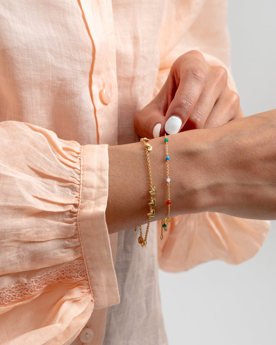 A model's wrist adorned with the Santa Monica Bracelet Gold, a delicate chain featuring multi-colored gemstones: Yellow Jade, Pink Opal, Sodalite, Green Onyx, Blue Color Reconstituted Turquoise, Vit Agate and Carnelian. Layered with a second gold bracelet with heart-shaped charms.