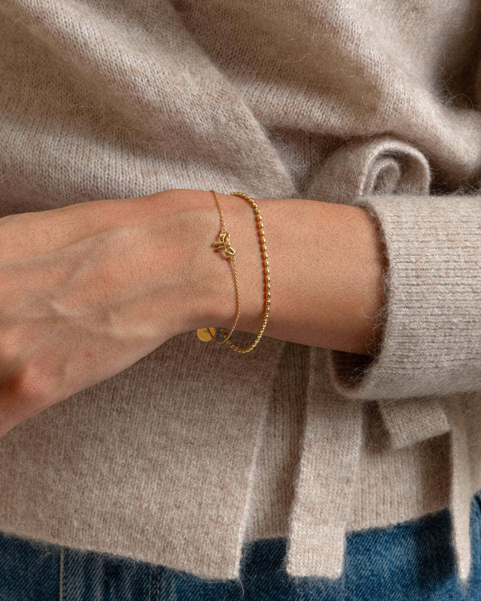 A close-up of a model wearing the Rosie Mini Bracelet in gold, which features a delicate chain with a small bow pendant and a second, structured chain.