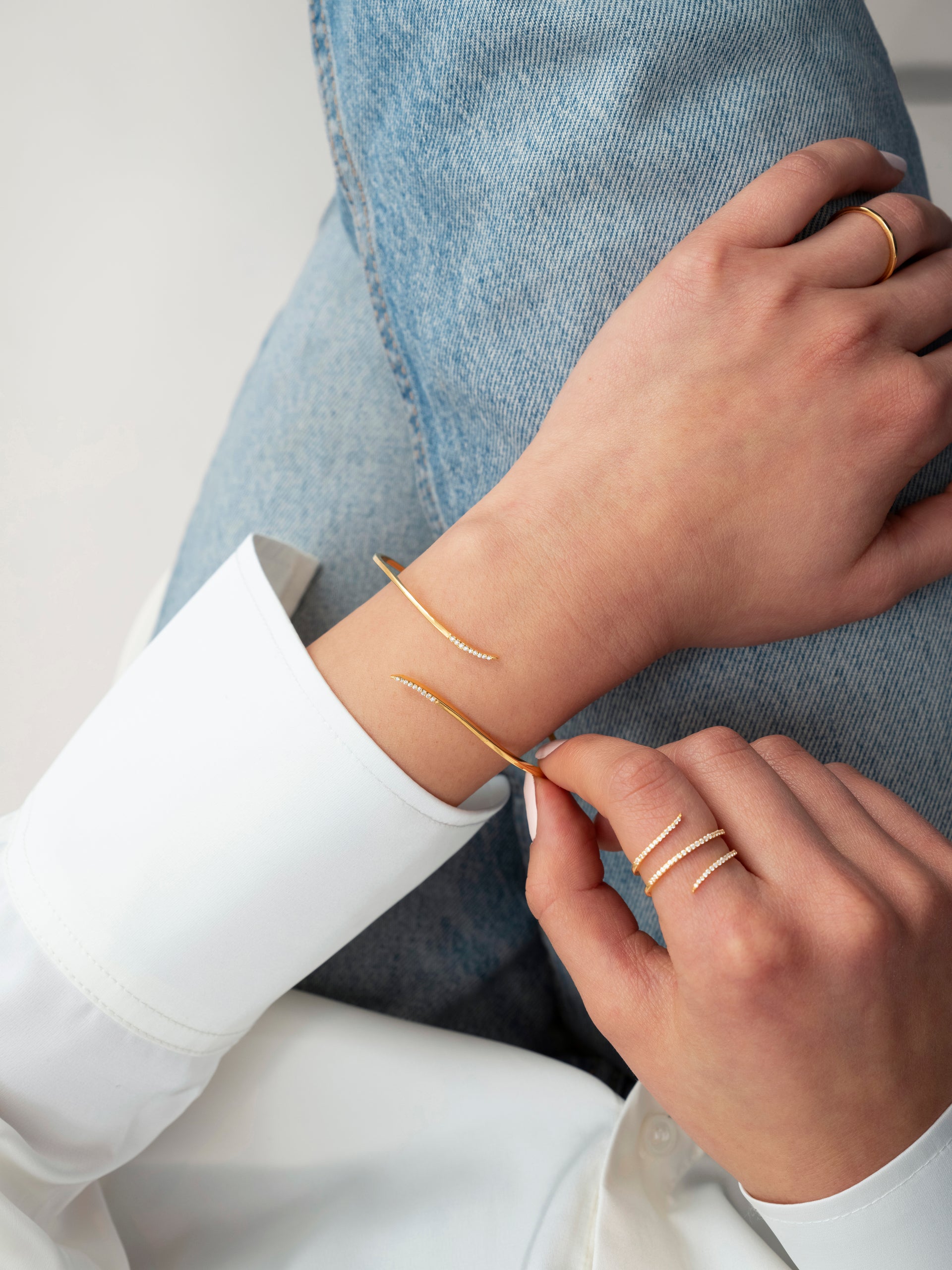 A model wearing the Sparkle Wrap Bangle in gold, which she is adjusting on her wrist. The bangle features curved ends paved with cubic zirconia. She is also wearing a matching Sparkle Wrap Ring and a simple gold band.