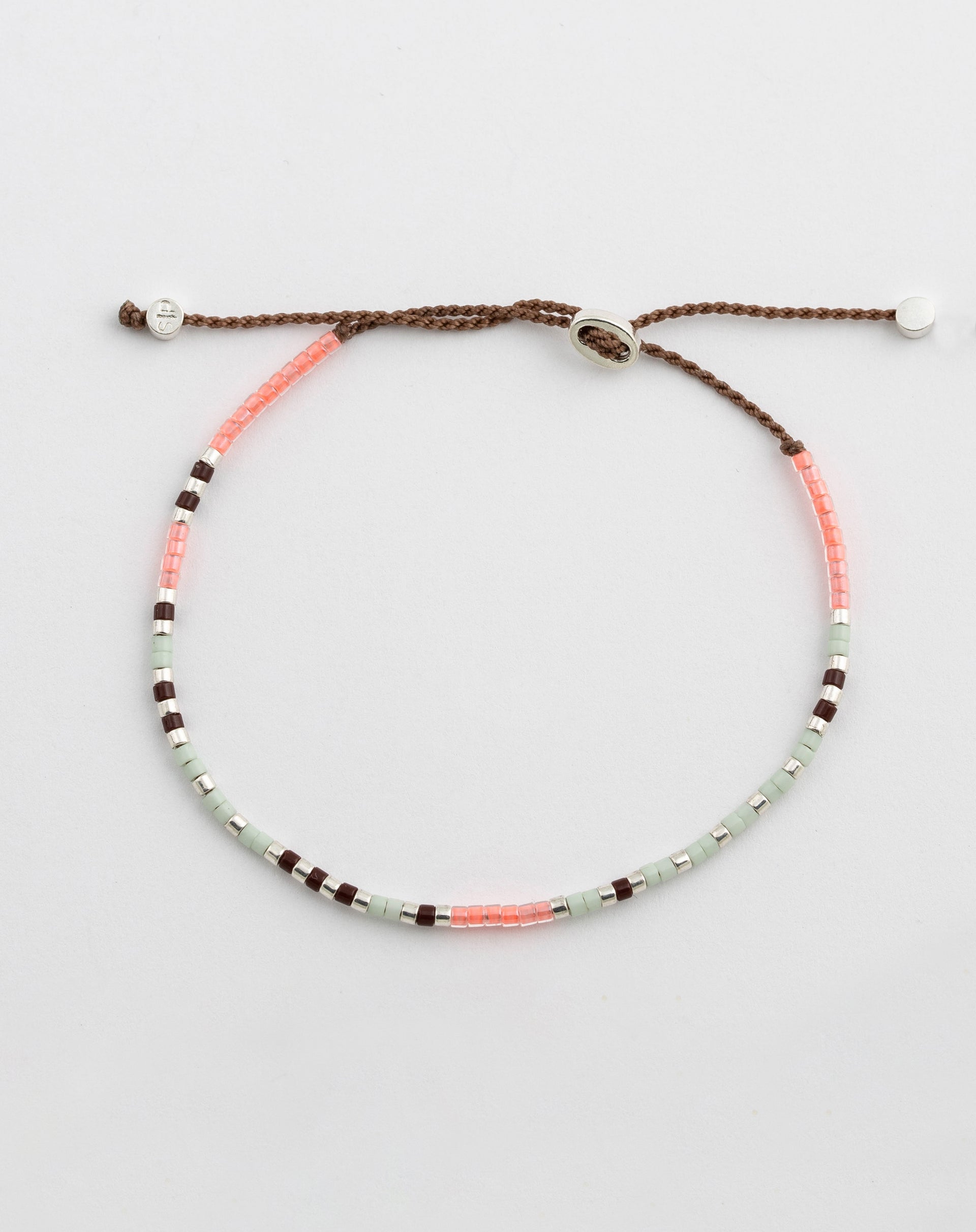 A product shot of the Code Bracelet Silver 'I Love You', laid out in a circle. It features pink, light green, and brown glass beads on an adjustable brown cord, arranged to spell 'I Love You' in morse code.