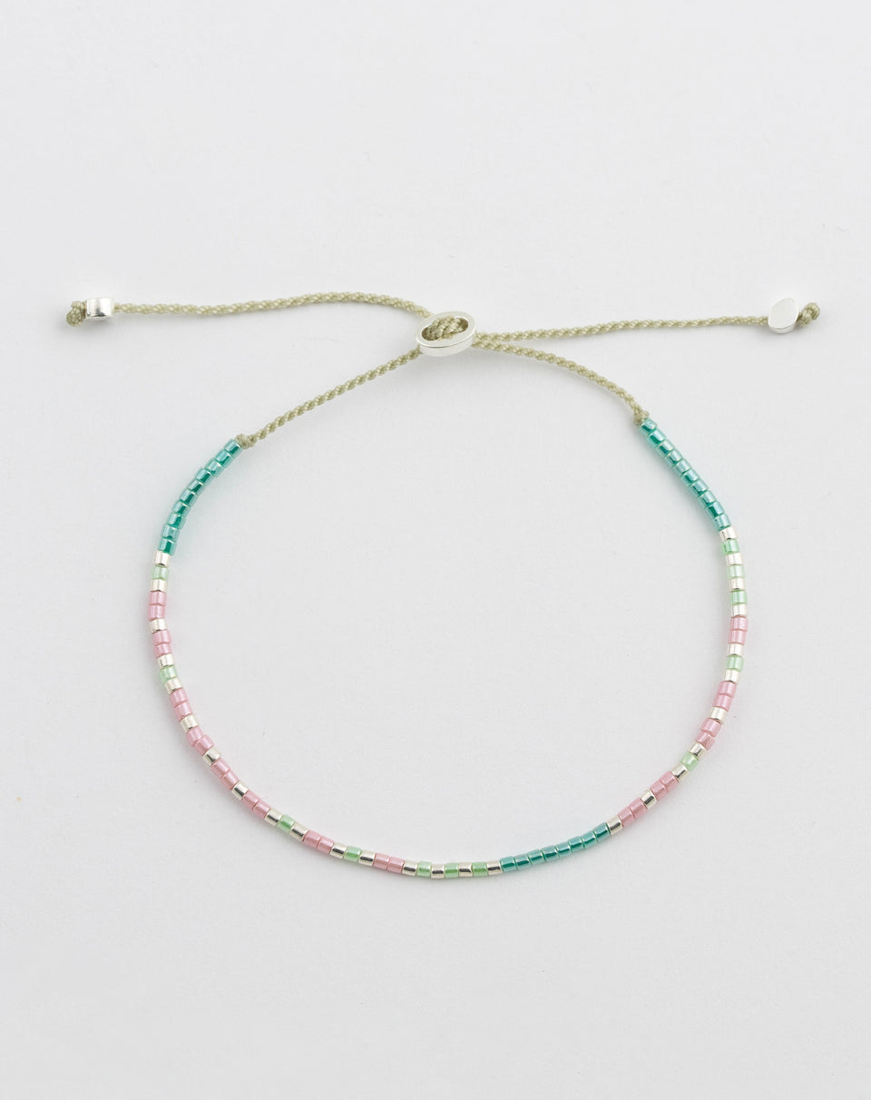 A product shot of the Code Bracelet Silver Stay Strong laid out in a circle. The bracelet features a delicate adjustable cord adorned with small Japanese glass beads in turquoise, pink, green, and silver, which spell out the message in Morse code.