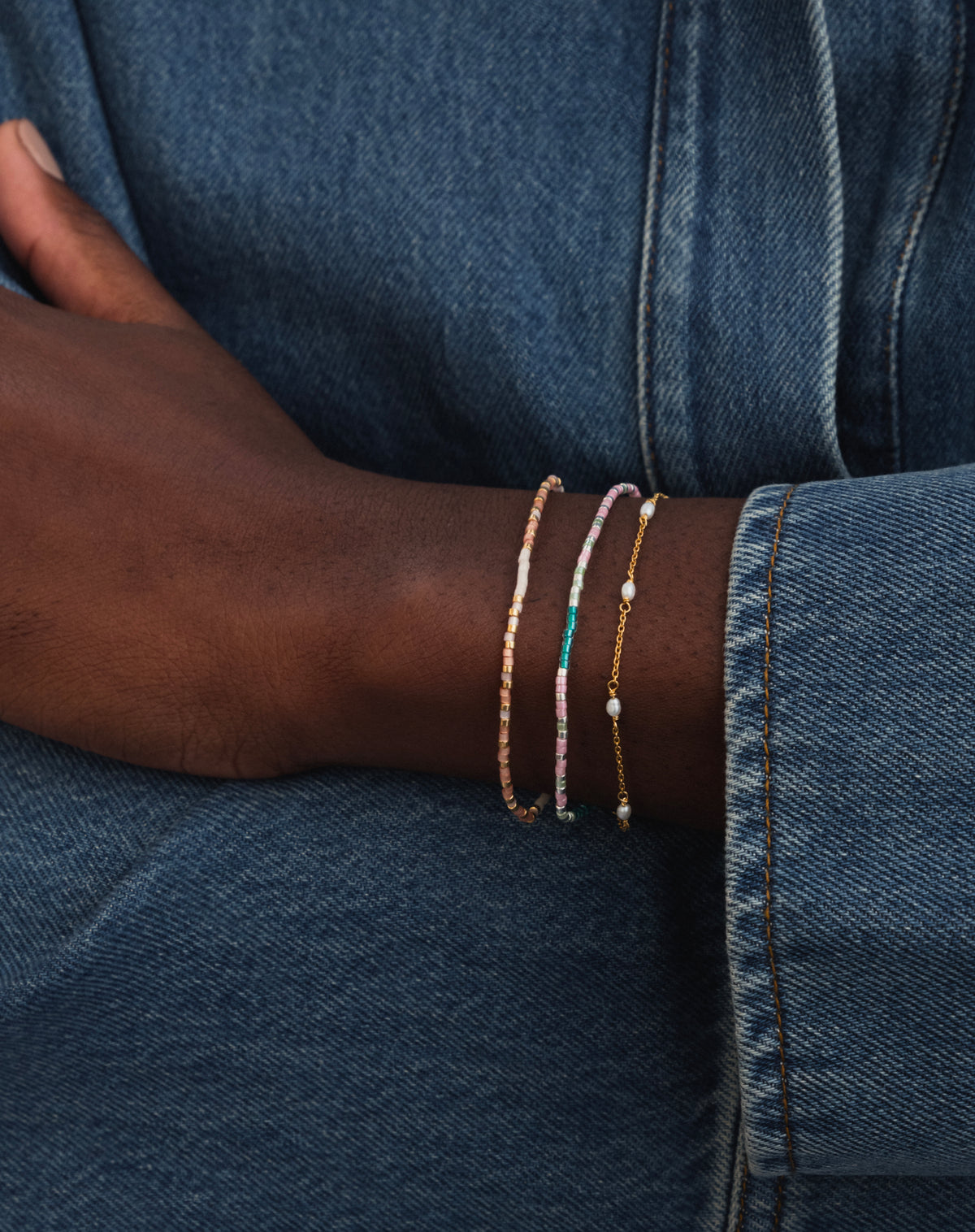 A model wears a stack of three Syster P bracelets against a denim background, featuring the 'Stay Strong' Code Bracelet with its pink, teal, and silver beads, a second beaded bracelet, and a fine gold chain with pearls.