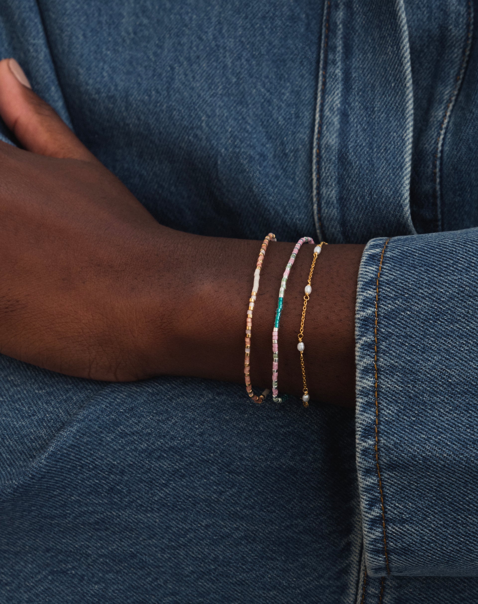 A model wears a stack of three Syster P bracelets against a denim background, featuring the 'Stay Strong' Code Bracelet with its pink, teal, and silver beads, a second beaded bracelet, and a fine gold chain with pearls.