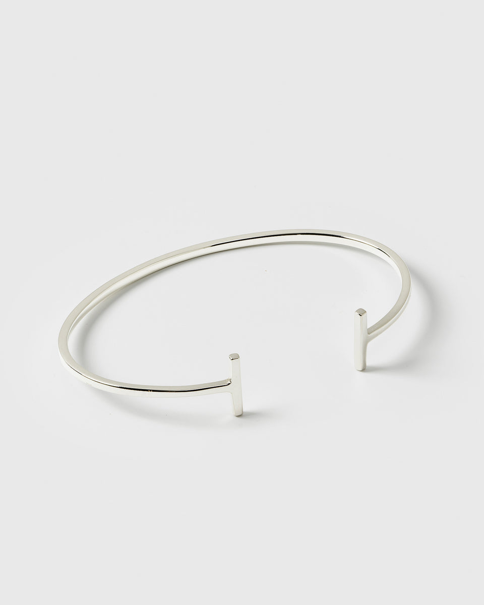 A product shot of the Strict Bar Bangle in silver, showing its thin, open design with a geometric bar at each end.