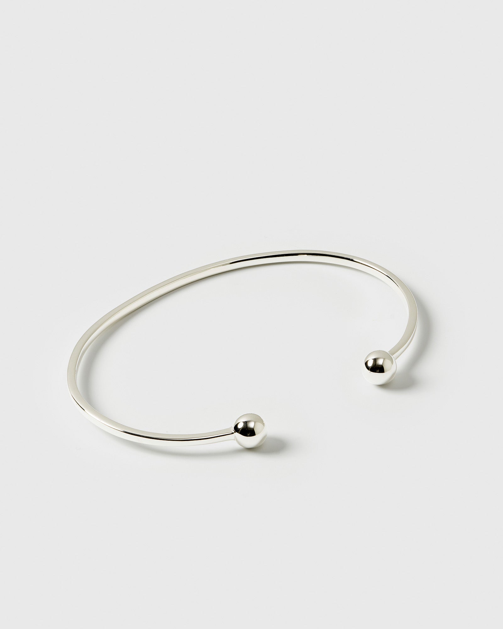 A product shot of the Strict Ball Bangle in silver, a thin, open cuff bracelet with a polished finish and small, rounded balls at each end.