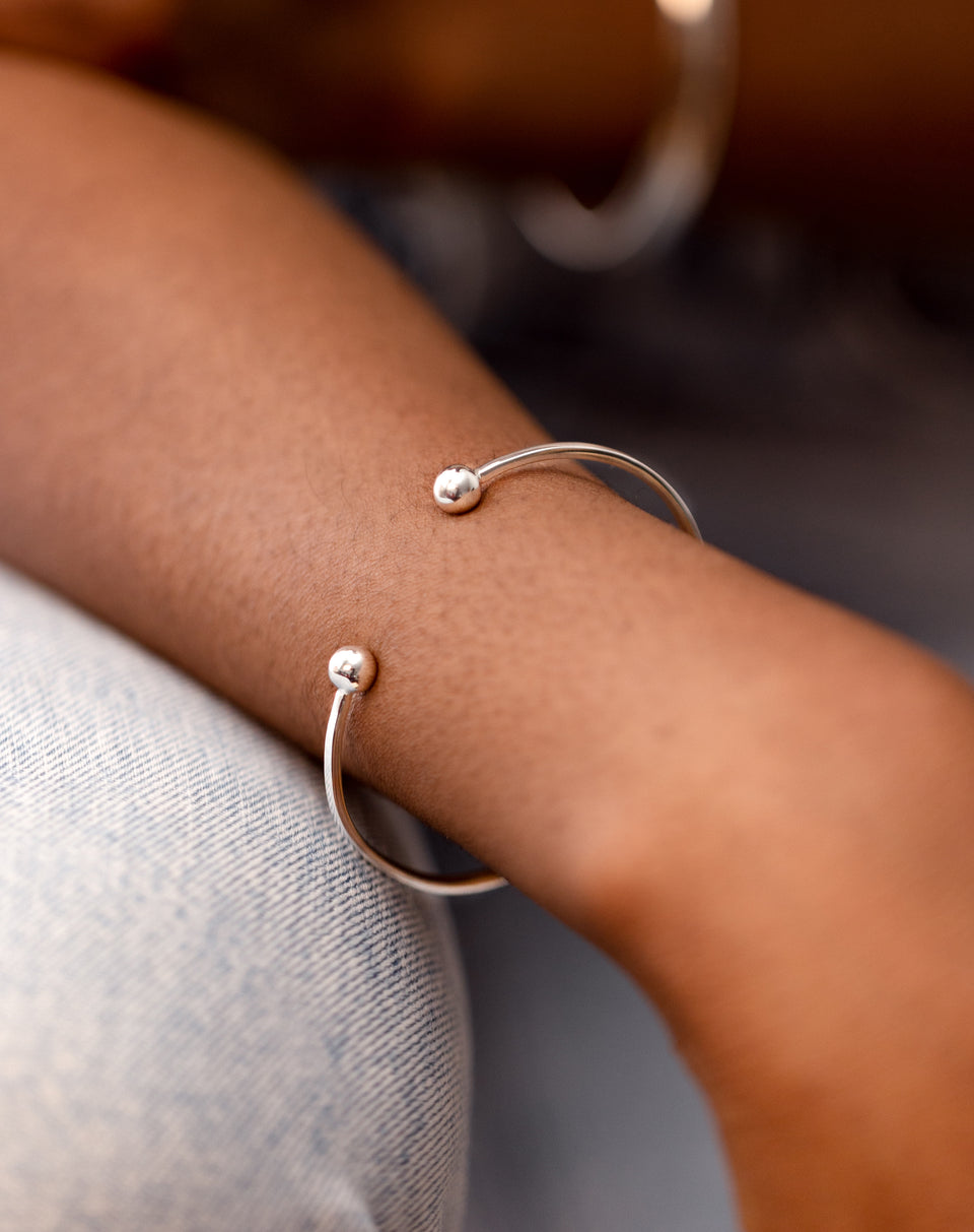 A close-up shot of a model wearing the Strict Ball Bangle in silver on her wrist, with a silver hoop earring slightly blurred in the background.