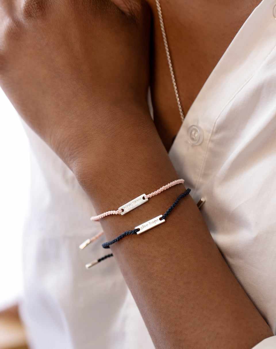 A close-up of a model wearing two Give Hope bracelets, one with a powder blue thread and another with a dark blue thread, each with a silver plaque engraved with 'Give Hope'.