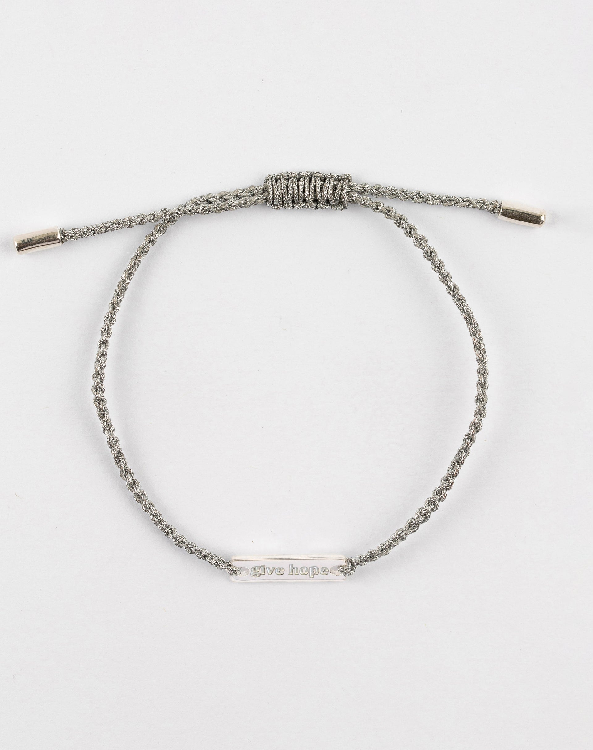 A product shot of the Give Hope bracelet in silver metallic, laid out in a circle. It features a braided metallic thread and a central silver plaque engraved with the words 'Give Hope'.