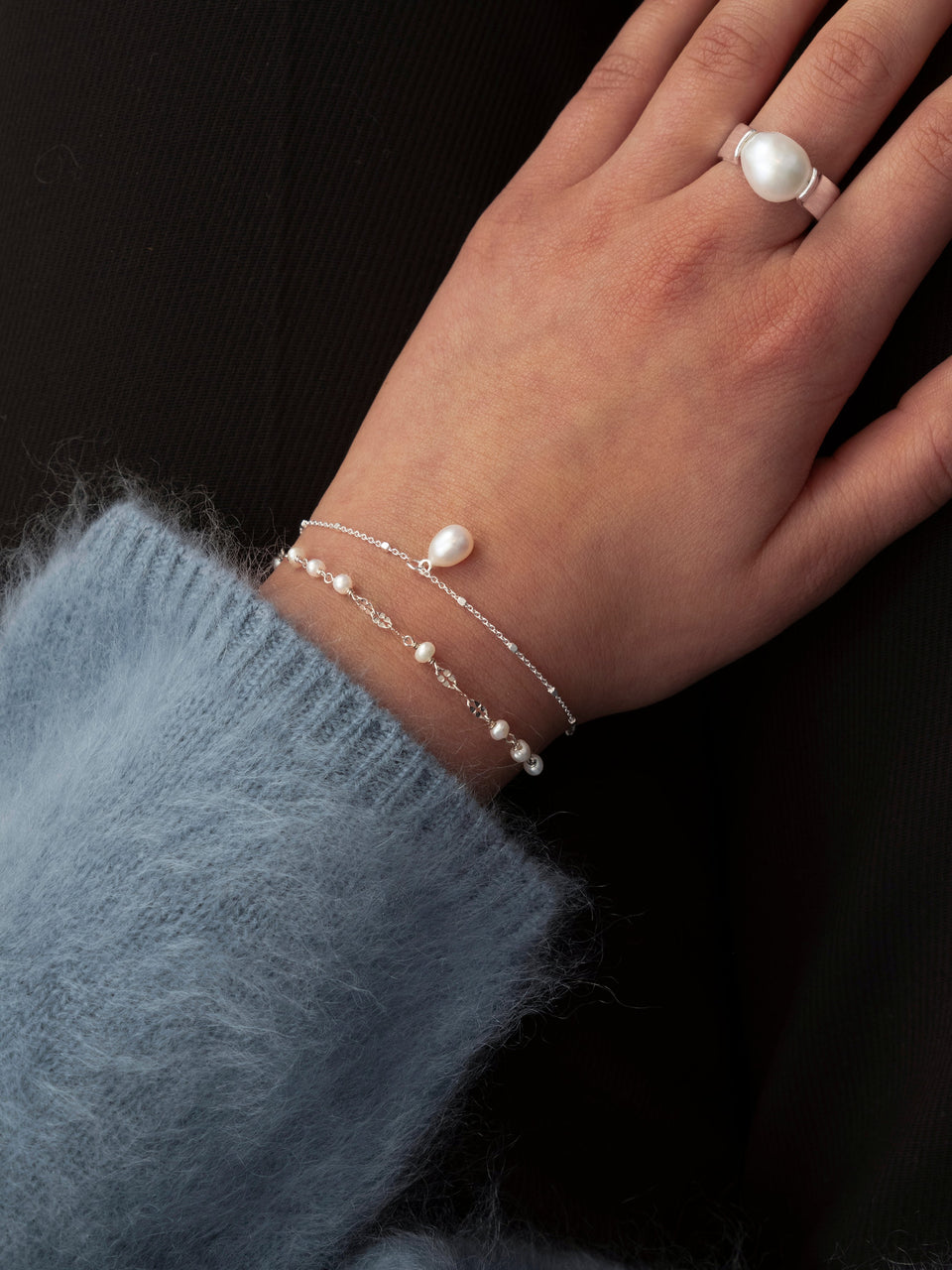 A close-up of a model's wrist wearing the Aurelia Bracelet in silver, which features a unique chain design with small freshwater pearls. She is also wearing a second delicate silver chain bracelet with a single pearl, and a wide silver ring set with a large pearl.
