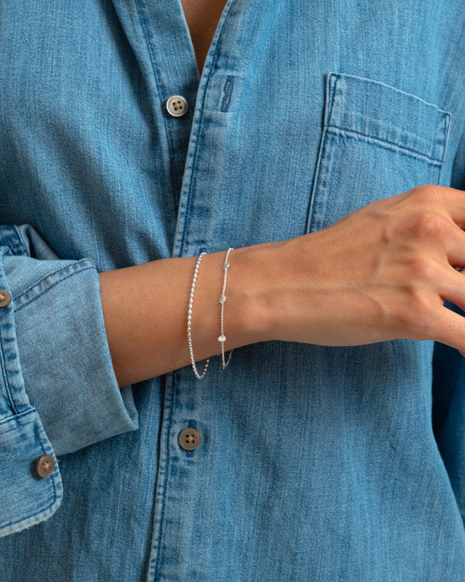 A model wears the delicate Solea Bracelet in silver, featuring small hammered circles, stacked with a simple silver beaded chain bracelet against a blue denim shirt.