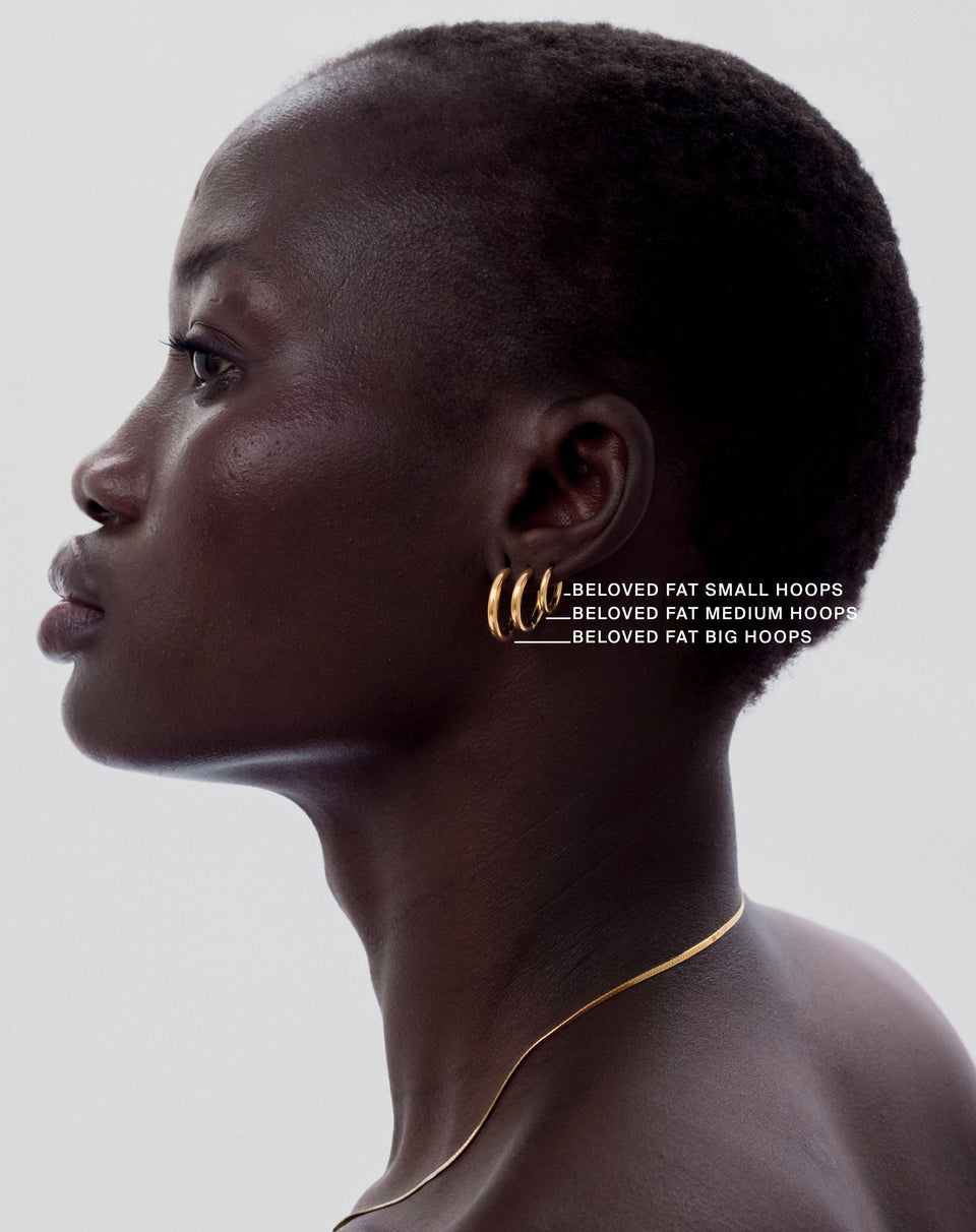 A model in profile wearing a stack of three gold Beloved Fat Hoops, with text labels identifying the small, medium, and big sizes, along with a fine gold chain necklace.