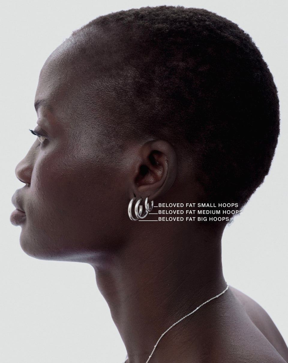 A size guide image showing a model in profile wearing the Beloved Fat Hoops in silver in three sizes‚ small, medium, and big‚ to illustrate their differences. She is also wearing a delicate silver chain necklace.