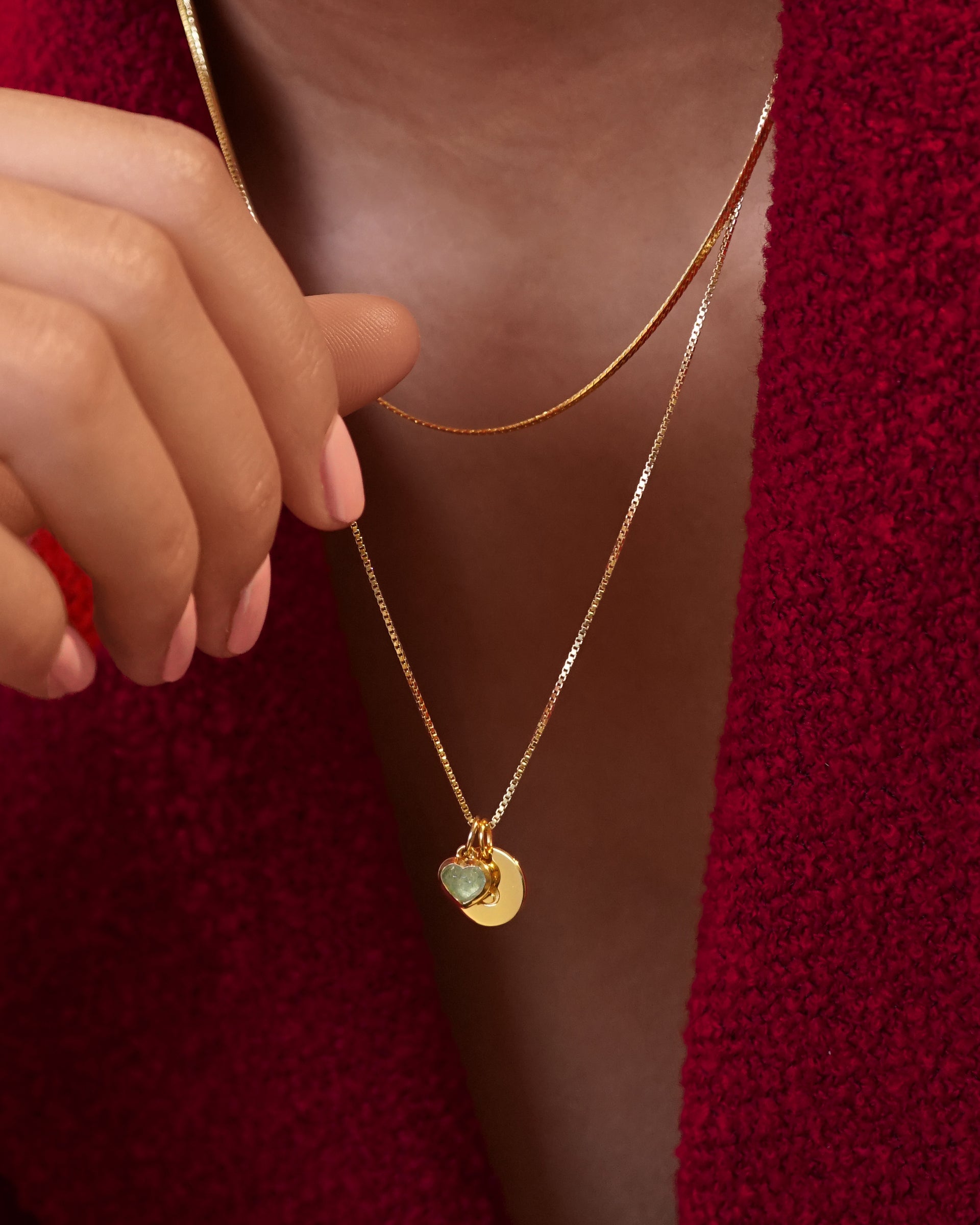 A model wears two layered gold necklaces against a red top: a flat snake chain and a finer chain with a green aventurine Beloved Heart Pendant and a round Beloved Mini Letter disc pendant.