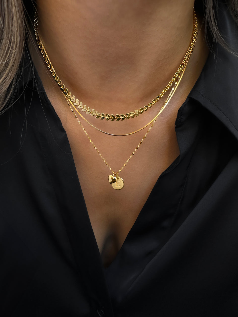 A close-up shot of a model wearing four layered gold necklaces, featuring a leaf-patterned chain, a flat snake chain, a simple delicate chain, and a longer chain with a round initial pendant and a heart charm.