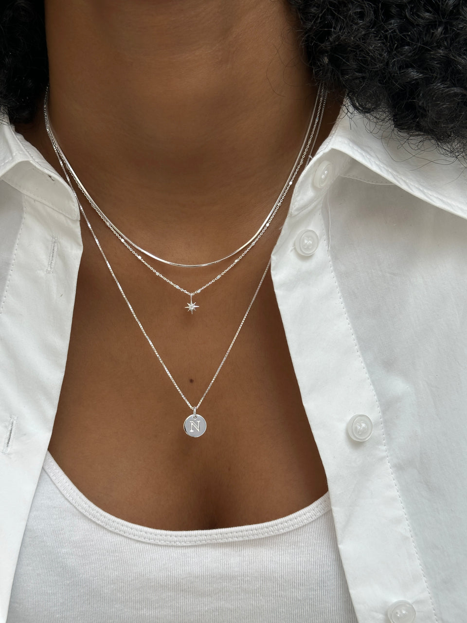 A close-up shot of a model wearing three layered silver necklaces: the Sheer Mirror Necklace, the North Star Necklace with a star pendant, and the Beloved Necklace with a circular 'N' initial pendant.