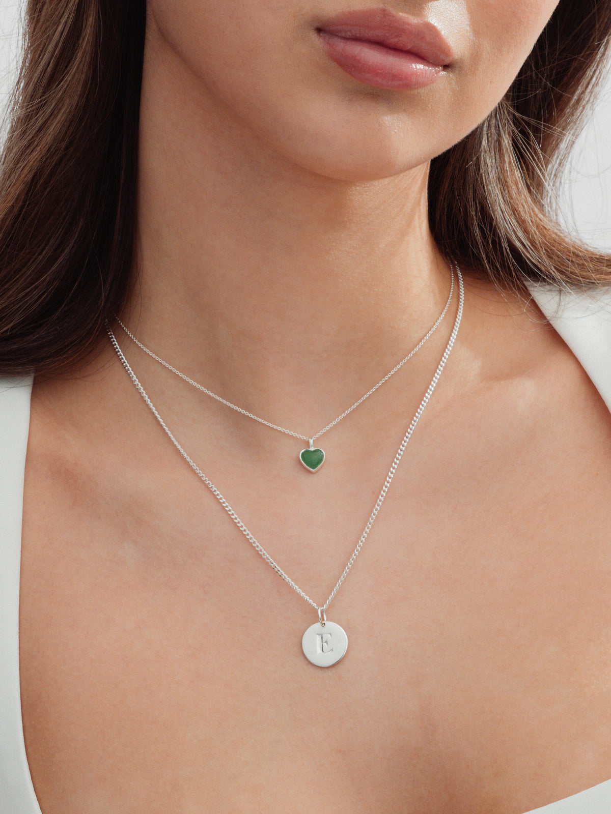 A close-up shot of a model wearing two layered silver necklaces: a shorter one with a green heart pendant and a longer one featuring the Beloved Short Curb Chain with a round letter 'E' pendant.