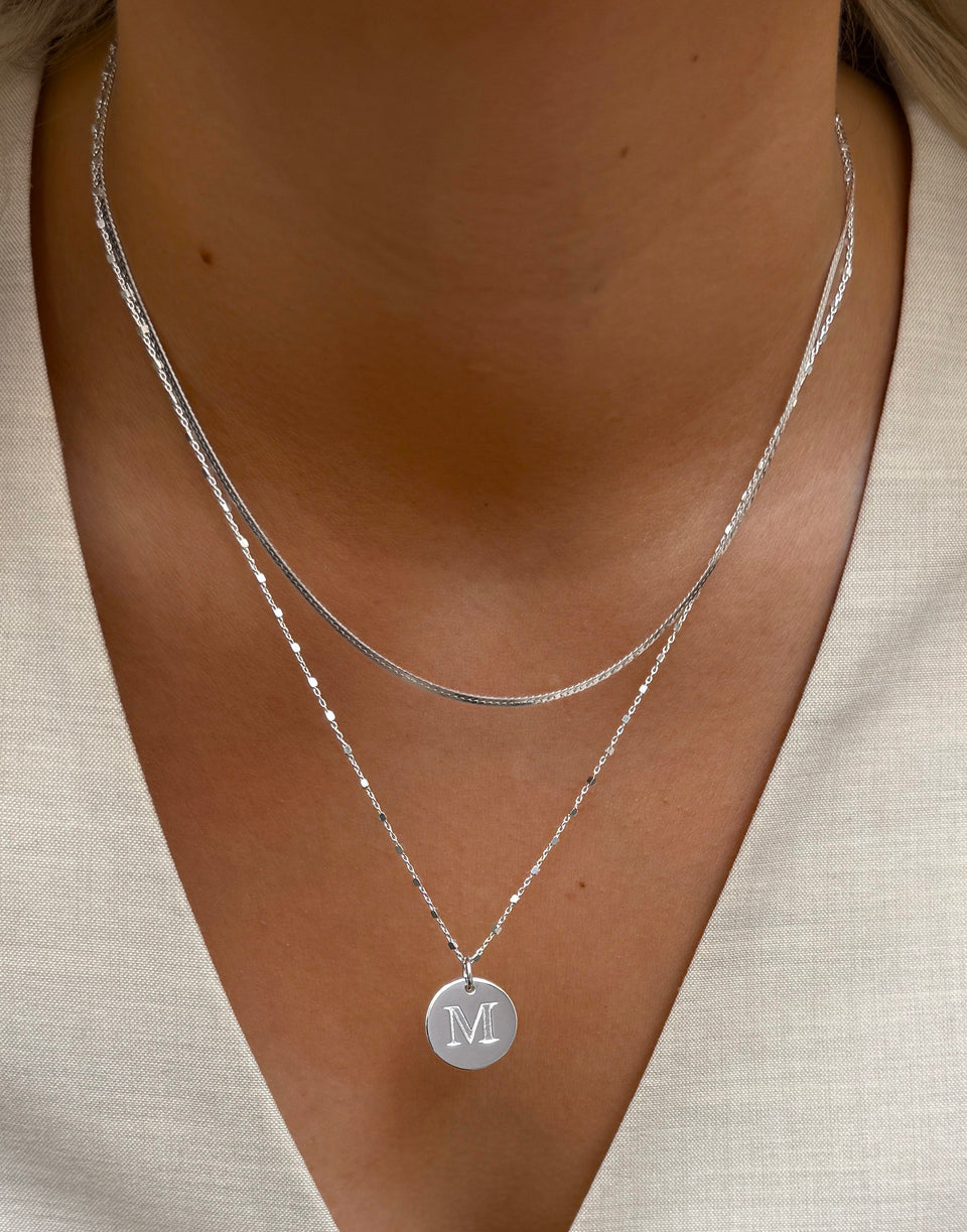 A close-up on a model's neck, showing the Beloved Big Letter Silver pendant with the letter 'M' on a delicate chain, layered with a shorter silver herringbone necklace.
