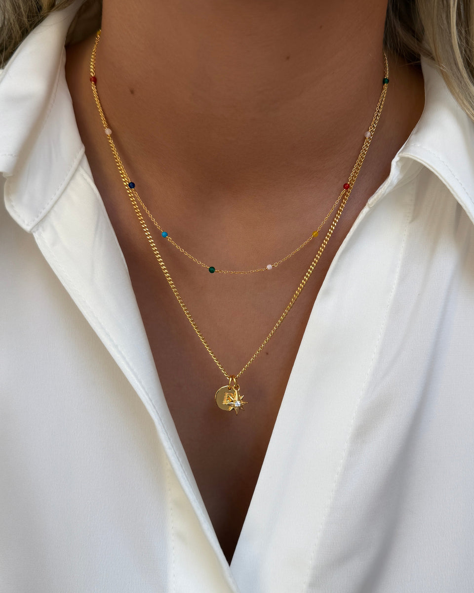 A close-up shot of a model wearing the Beloved Compass Star Pendant in gold on a chain, paired with a round initial pendant. The necklace is layered with a second, finer gold chain featuring small, multi-colored beads.