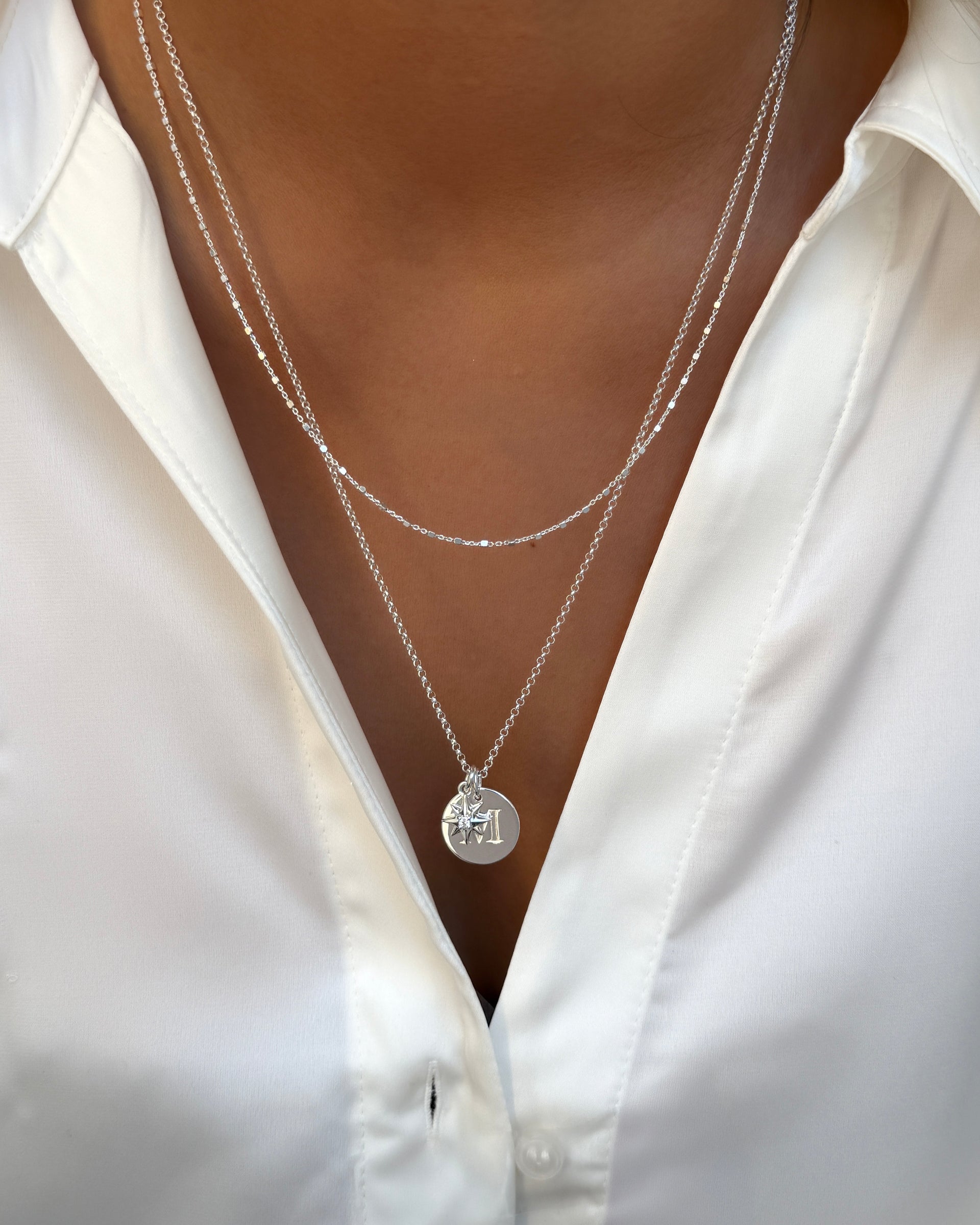 A model wearing the Beloved Compass Star Pendant in silver, styled on a chain with a round 'M' initial pendant and layered with a second, more delicate silver chain.
