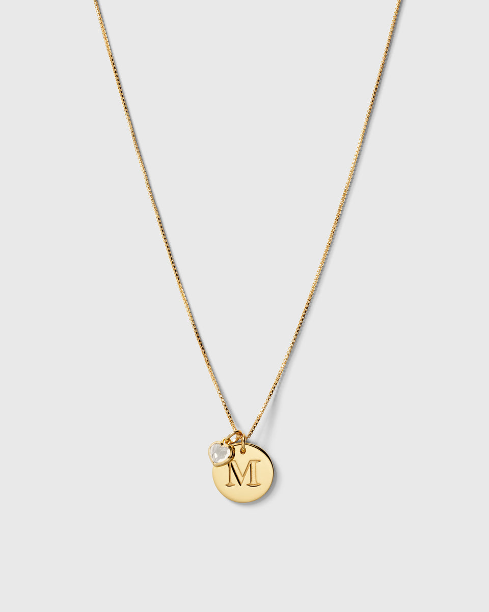 A product shot of the gold Beloved Heart Pendant, styled on a delicate box chain with a round initial 'M' pendant.