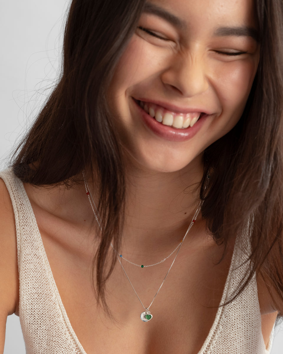A smiling model wearing a layered silver necklace set, featuring the Beloved Heart Pendant with a green aventurine stone and a round silver disc pendant, styled with a shorter, delicate beaded necklace.
