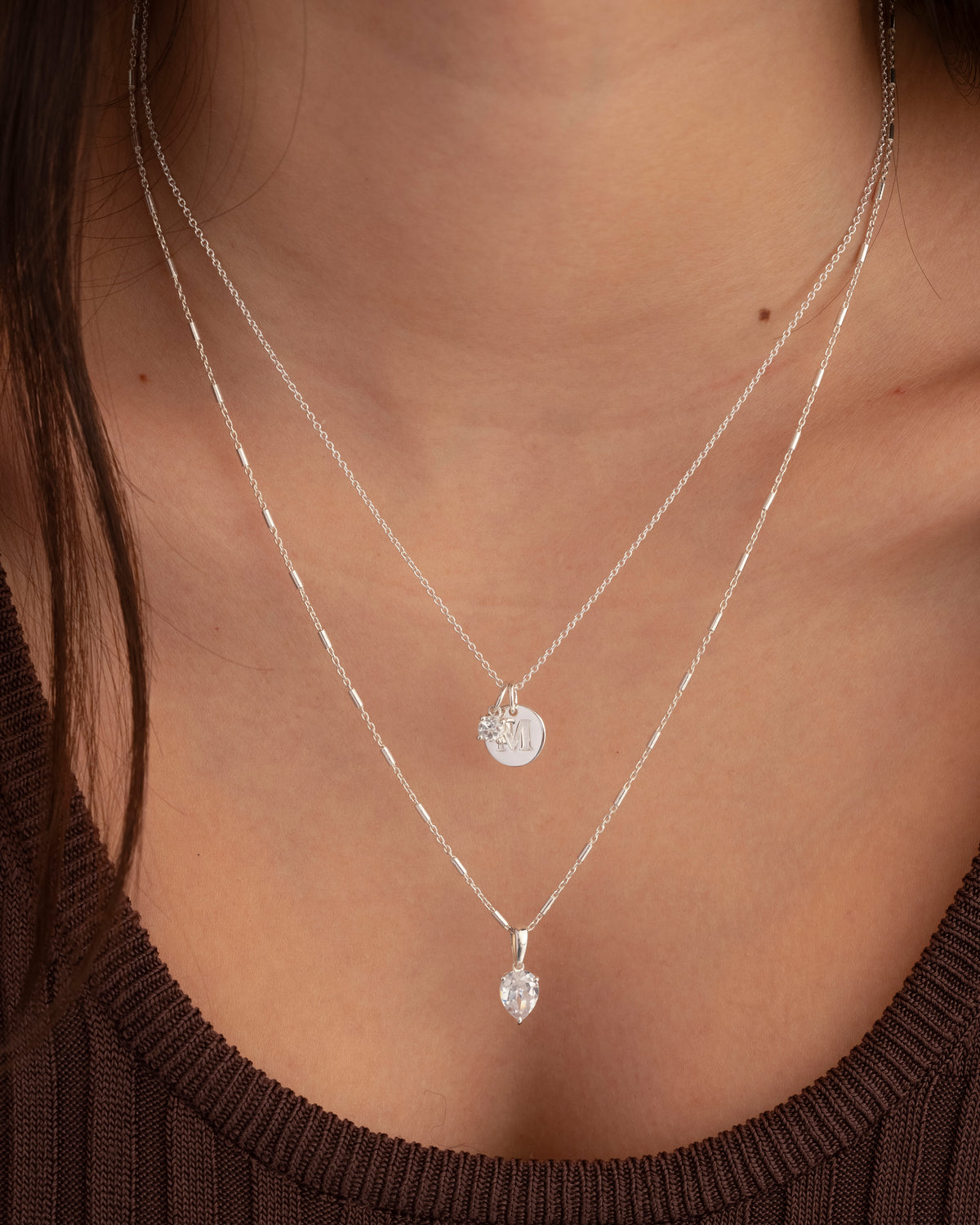 A close-up shot of a model's neck, showing two layered silver necklaces. The longer necklace features the Beloved Solo Nova Pendant with its teardrop-shaped white topaz, while the shorter necklace has a small, round initial pendant and a clear stone charm.