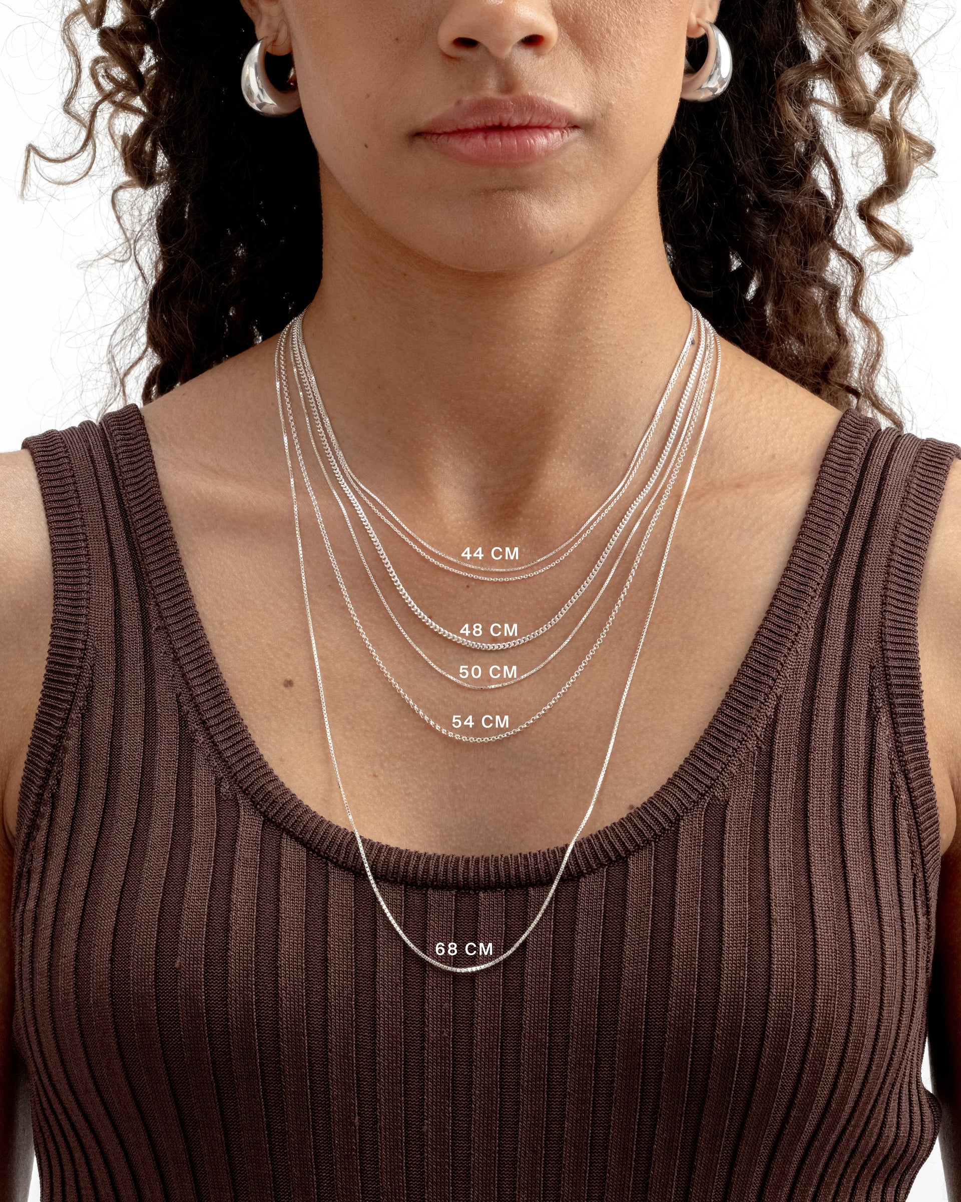 A size guide for Syster P silver necklaces, showing a model wearing five layered chains of different styles to demonstrate lengths from 44cm to 68cm, also wearing a pair of chunky silver hoop earrings.