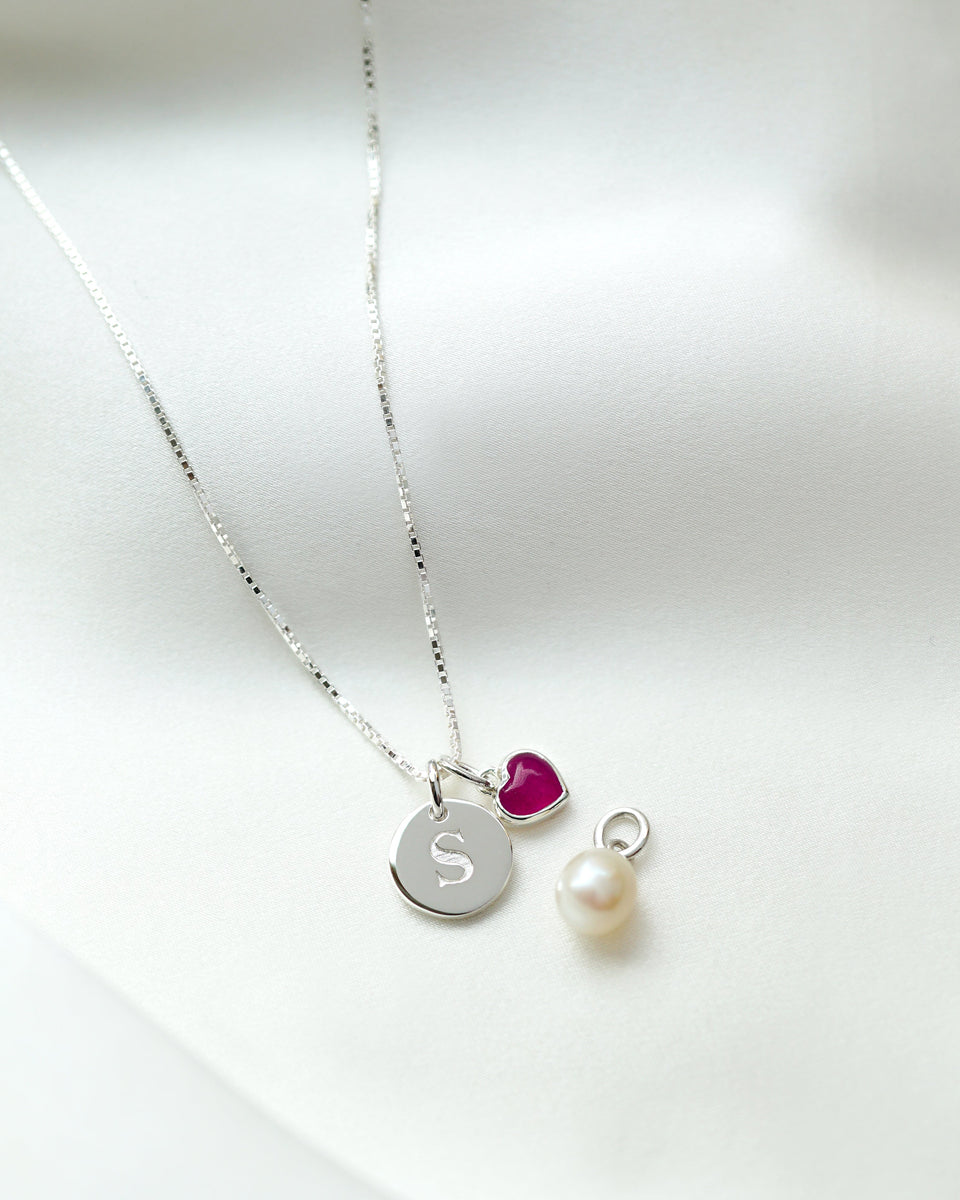 Silver box chain necklace with two pendants: a round letter and a heart-shaped pink quartz, alongside a single pearl pendant.