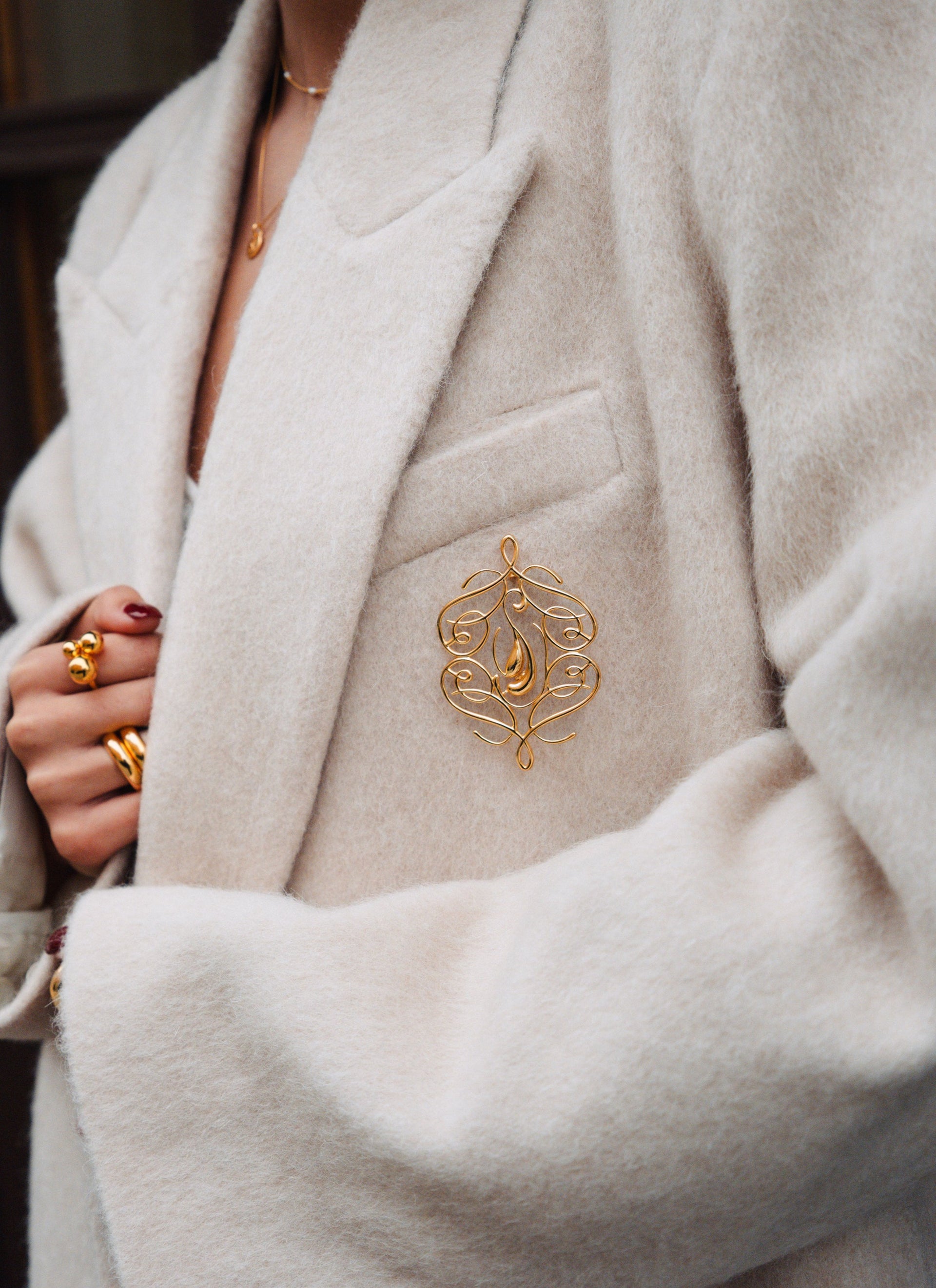 A model styles the gold Ophelia Brooch on the lapel of a cream-colored coat, complemented by the gold Universe Ring and Bolded Ring on her hand, and a delicate gold necklace.