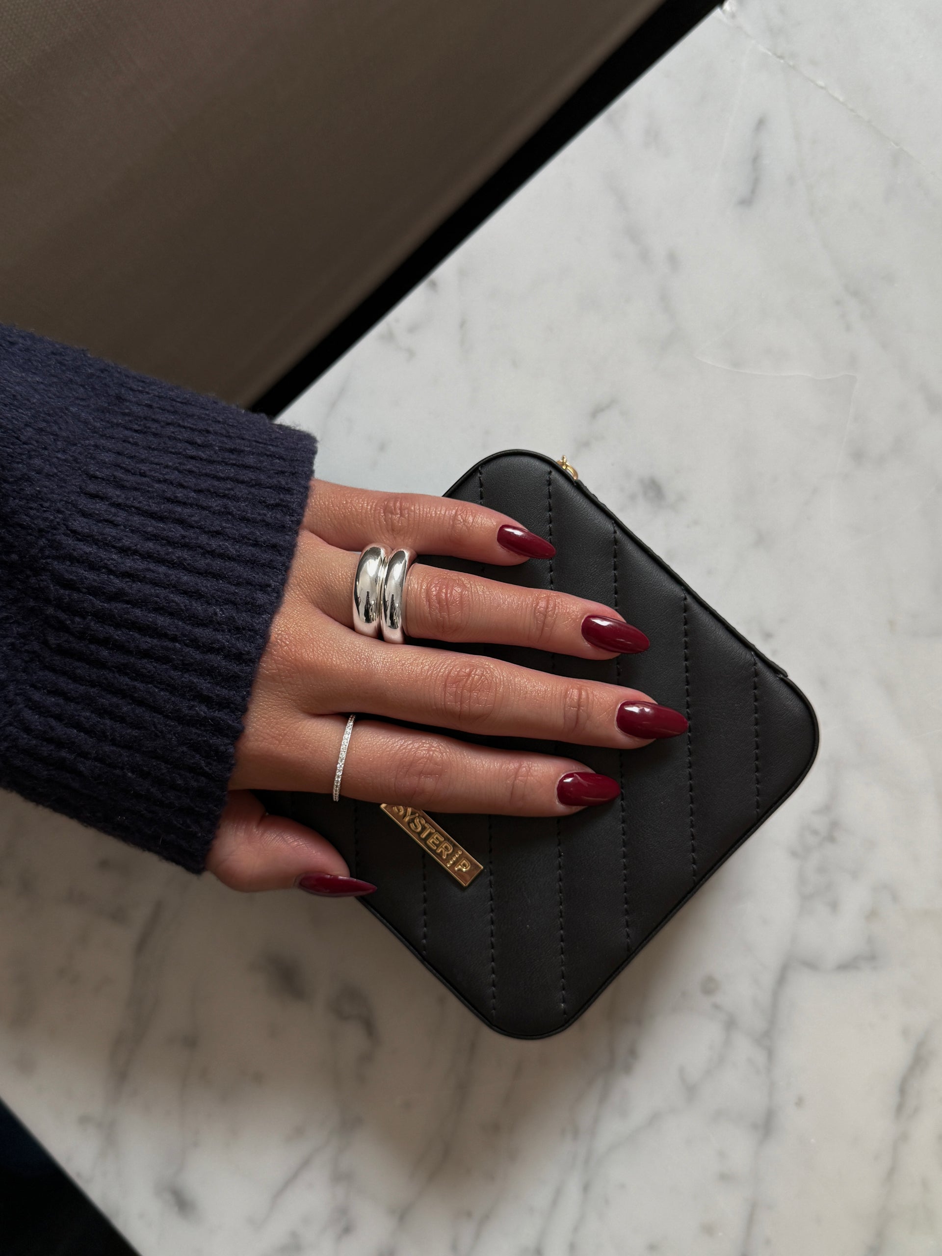 A model's hand, wearing a bold double-band silver ring and a thin sparkling pavé band, rests upon the small black quilted Syster P jewelry case.